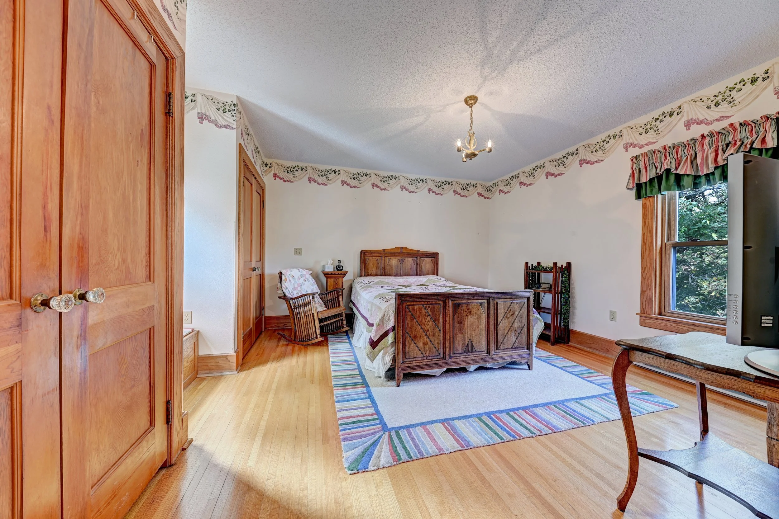 A bedroom with wooden flooring and wooden furniture, including a wardrobe, bed, and chairs. The room has a colorful striped rug and window valances with a floral and pink design. A ceiling chandelier hangs from the ceiling, and there is a television 