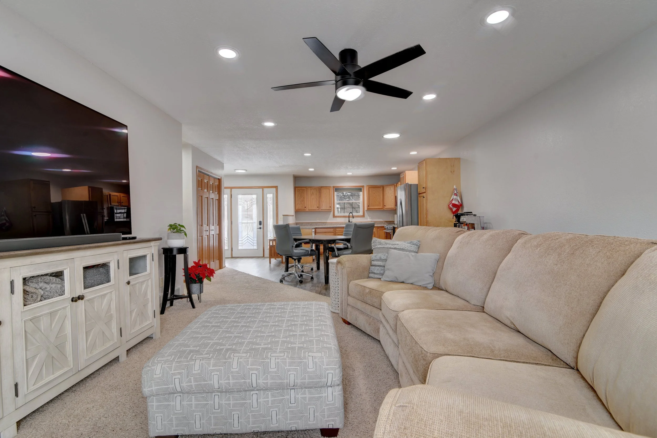 Spacious open-concept living room with large beige sectional sofa, patterned ottoman, entertainment center, and a view into the kitchen with wooden cabinets and a dining area with chairs, ceiling fan, and recessed lighting.
