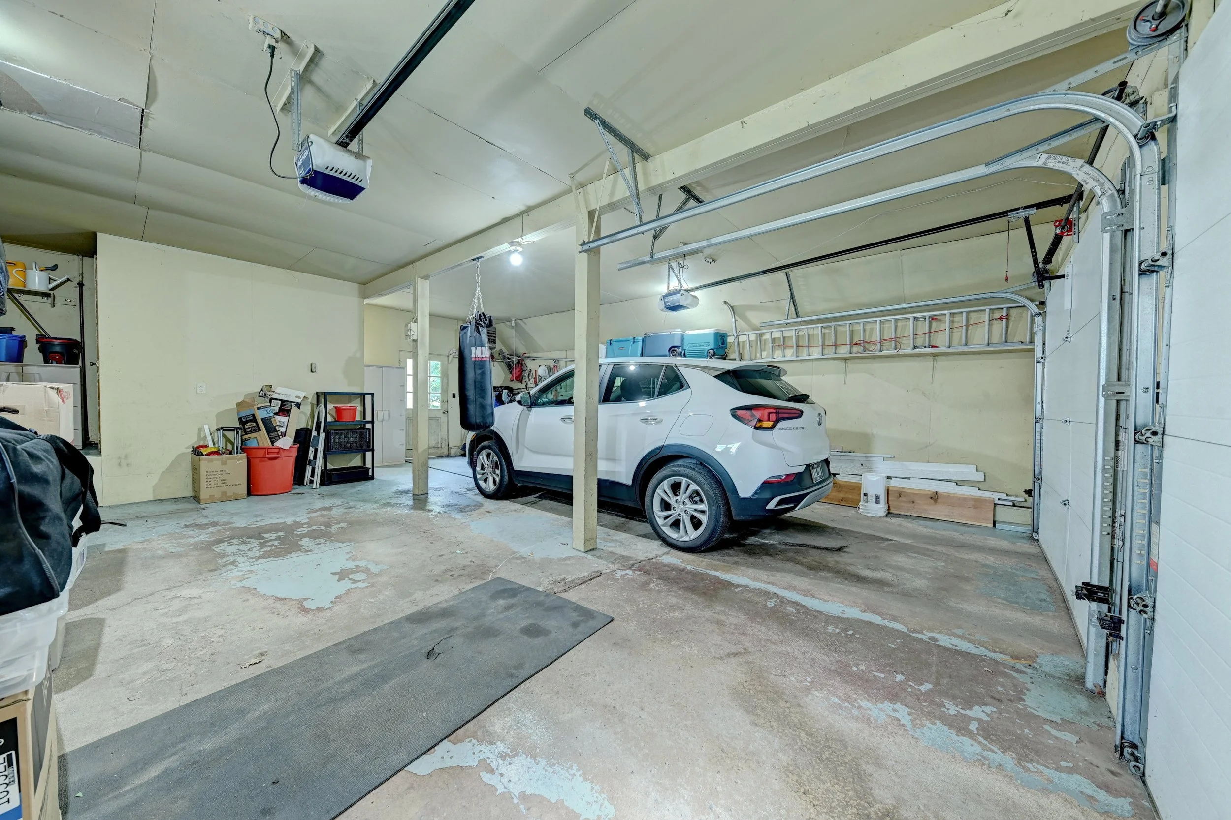 A spacious garage with a white car parked inside. The garage has a concrete floor with some peeling paint, a hanging punching bag, shelves with various items, and a garage door opener system on the ceiling.