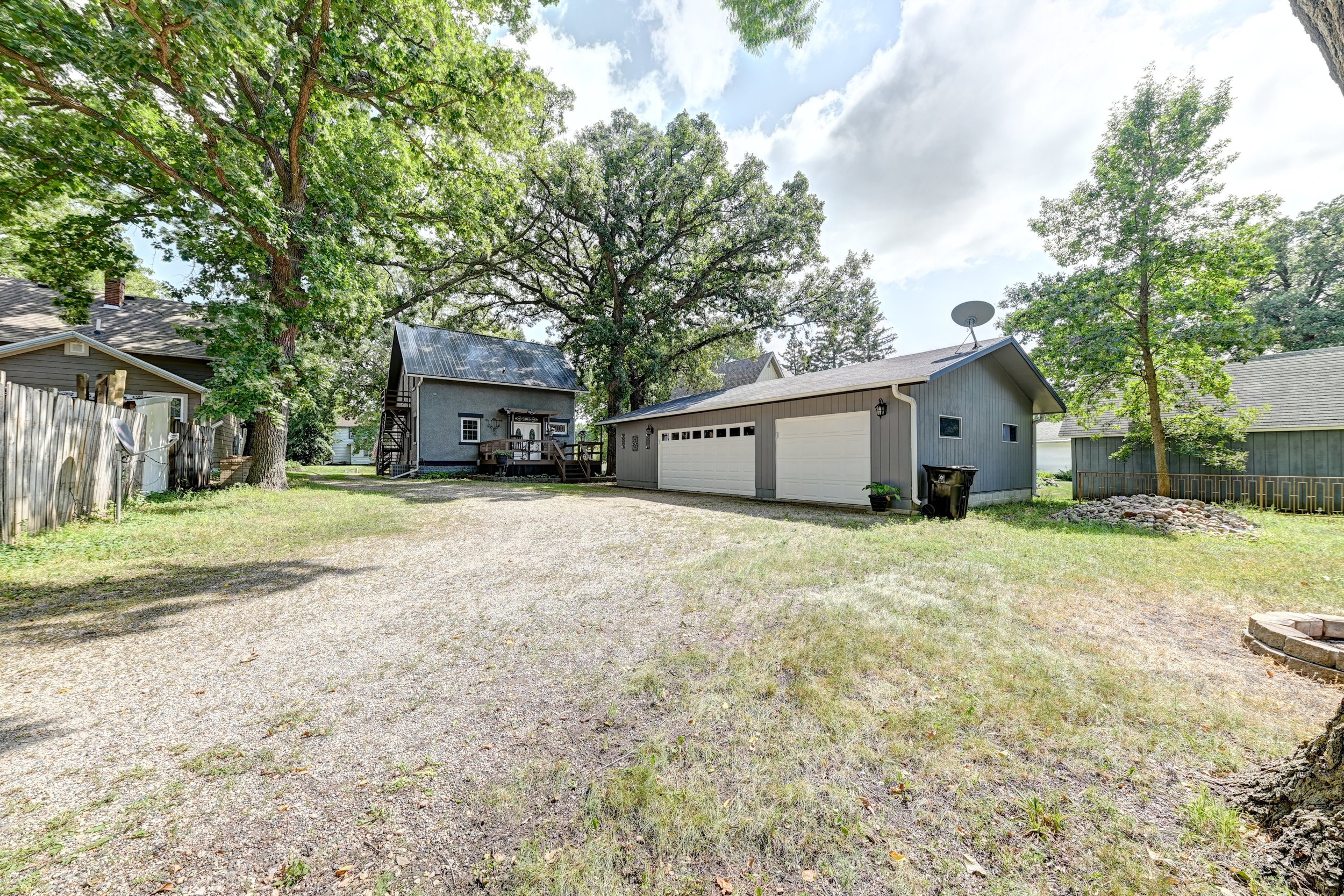 A backyard with a large gravel driveway, two trees, a grey garage with a white door, a small grey building with a metal roof, and a wooden shed. There is also a satellite dish on the roof of the garage.