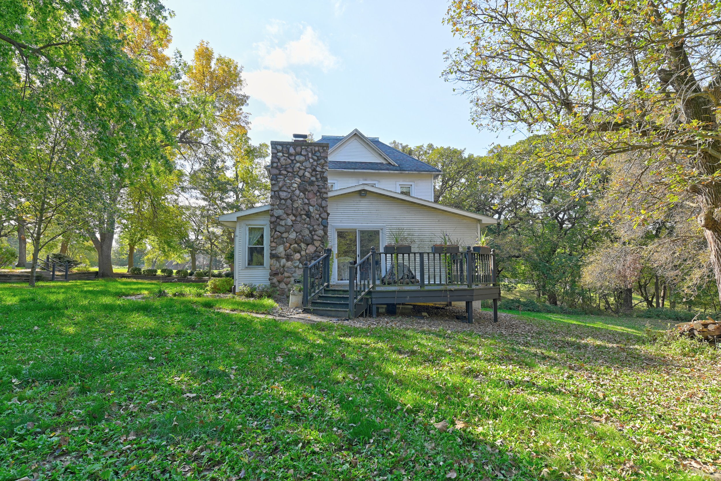 A white house with a stone chimney and a wooden deck, surrounded by green grass and trees with some autumn leaves, under a partly cloudy sky.