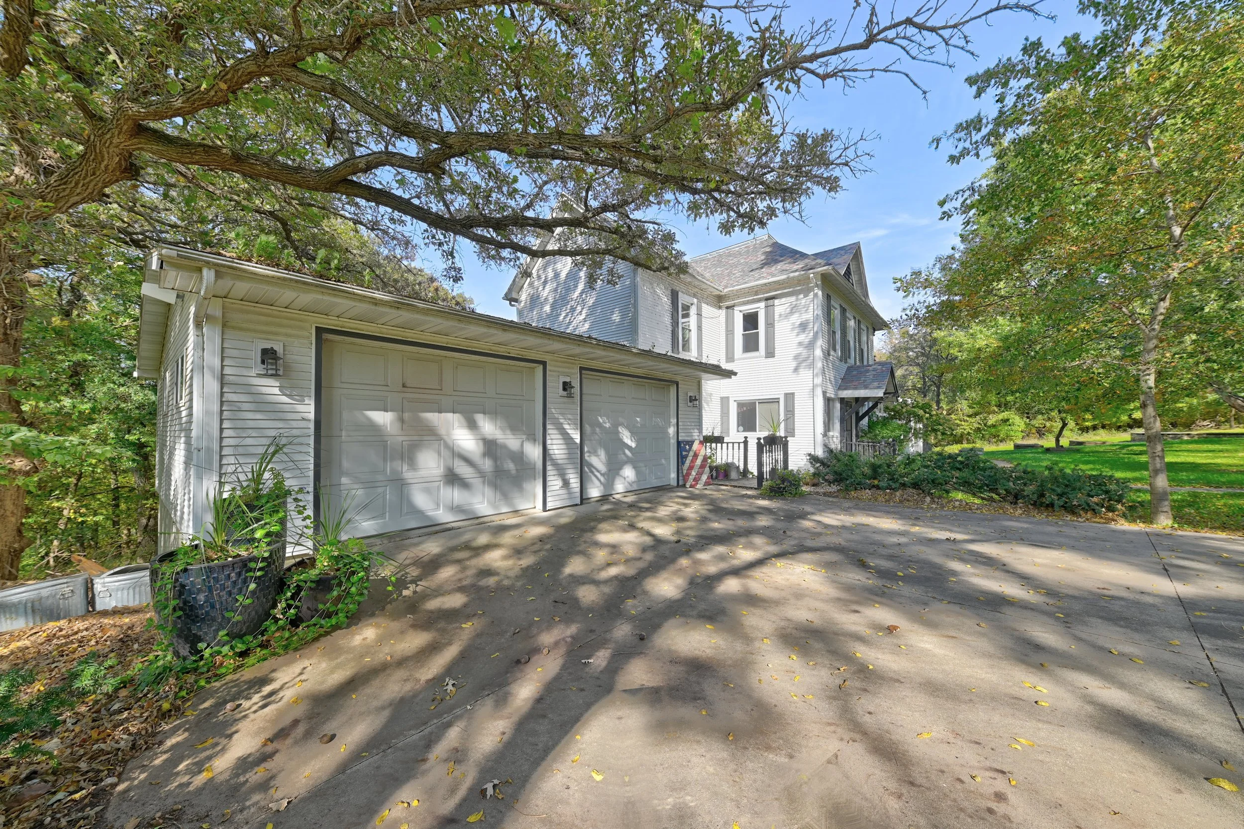 A white two-story house with a detached garage, surrounded by green trees and a yard, under a blue sky with some clouds.