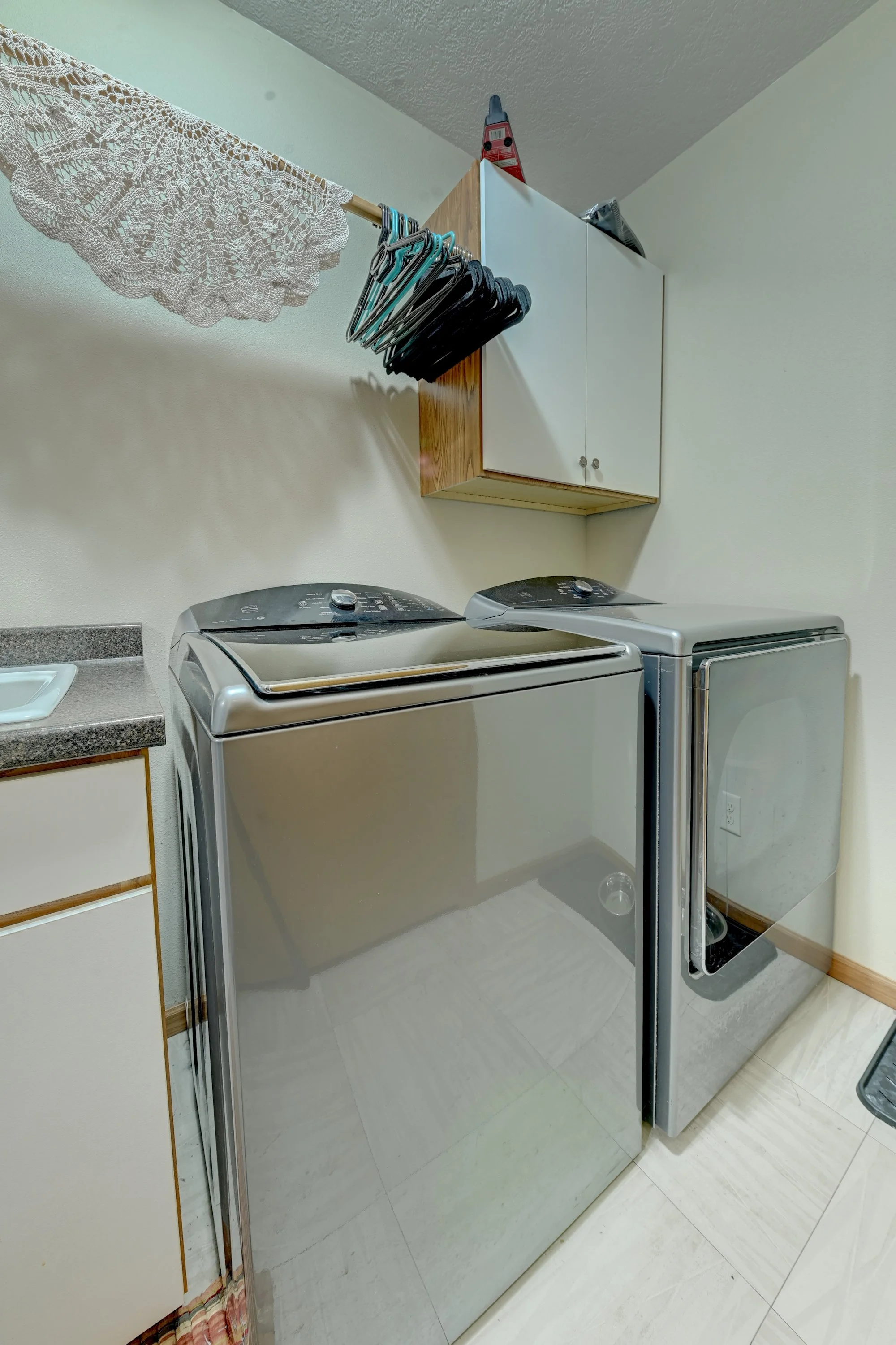 A laundry room with a top-loading washing machine and a side-by-side dryer, white cabinets, some hangers on a rack, and a lace tablecloth hanging on a drying rod.