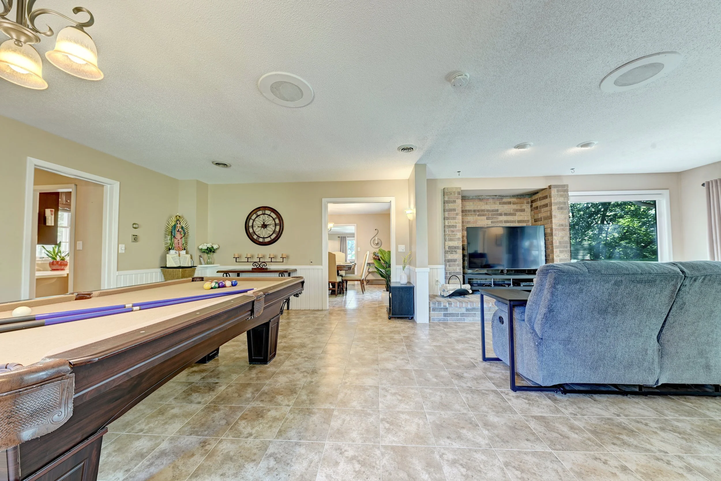 Living room with a pool table in the foreground, a large flat-screen TV on a brick wall, a blue couch, a dining area with a table and chairs, and large windows showing greenery outside.