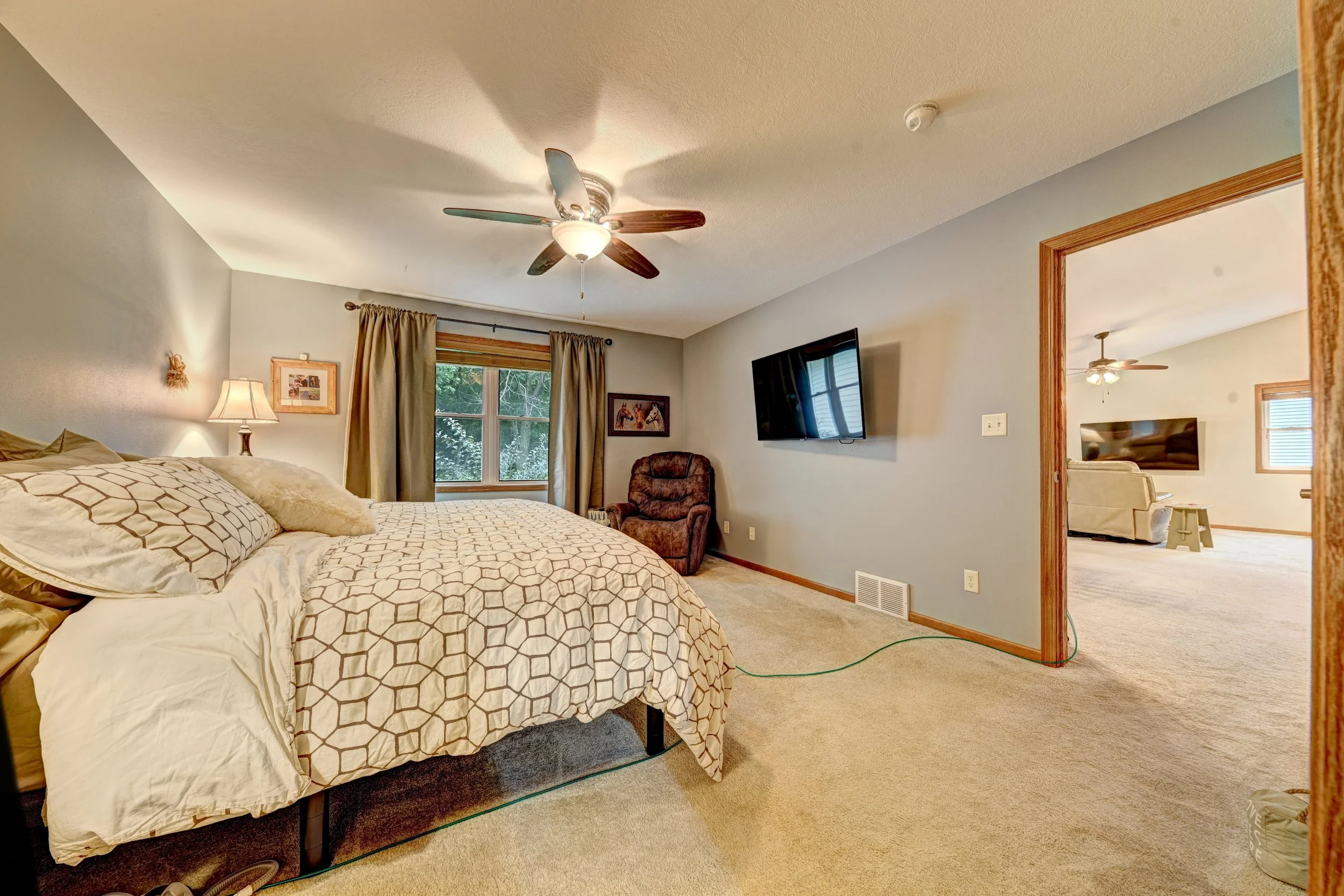 A bedroom with a bed, patterned bedding, a bedside lamp, a window with curtains, a wall-mounted TV, a chair, and an open doorway leading to another room. The room has beige walls and carpeted flooring.