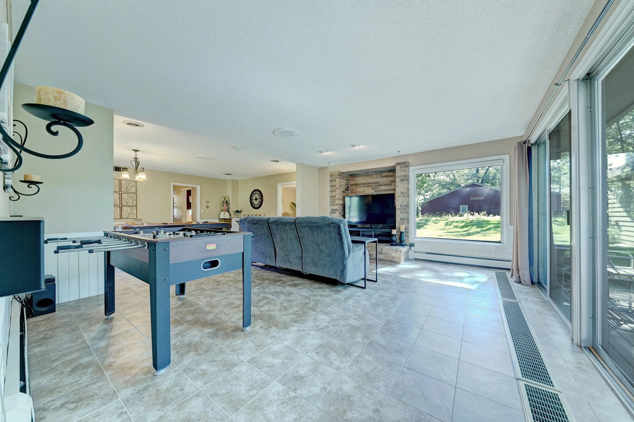 Living room with tile flooring, a blue foosball table, a gray sofa, a brick fireplace, large windows with views of trees outside, and various decorations including a wall clock and candles.