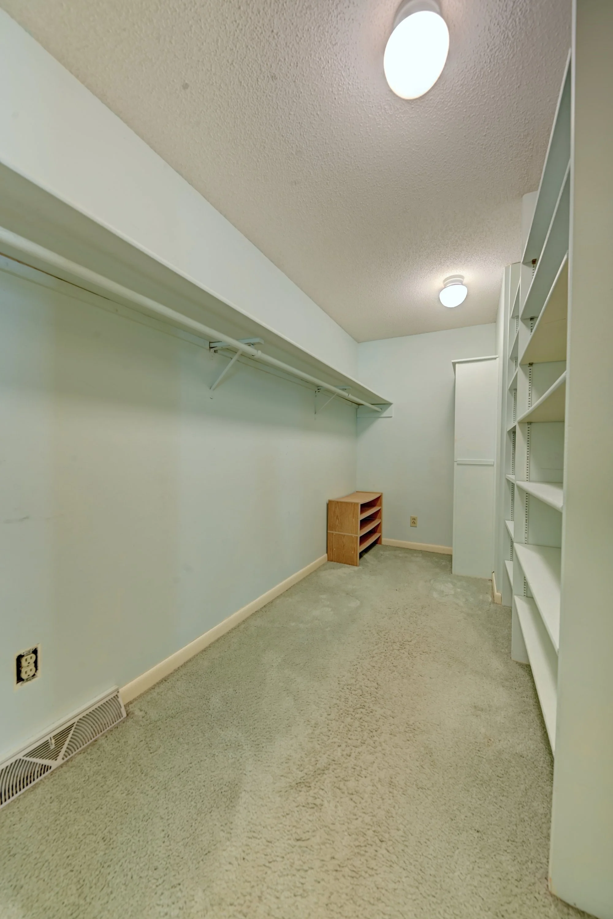 Empty walk-in closet with built-in white shelves on the right and a clothing rod on the left. The walls are painted light blue, and the floor is carpeted. Two ceiling lights are turned on, and a small wooden shoe rack is visible in the corner.