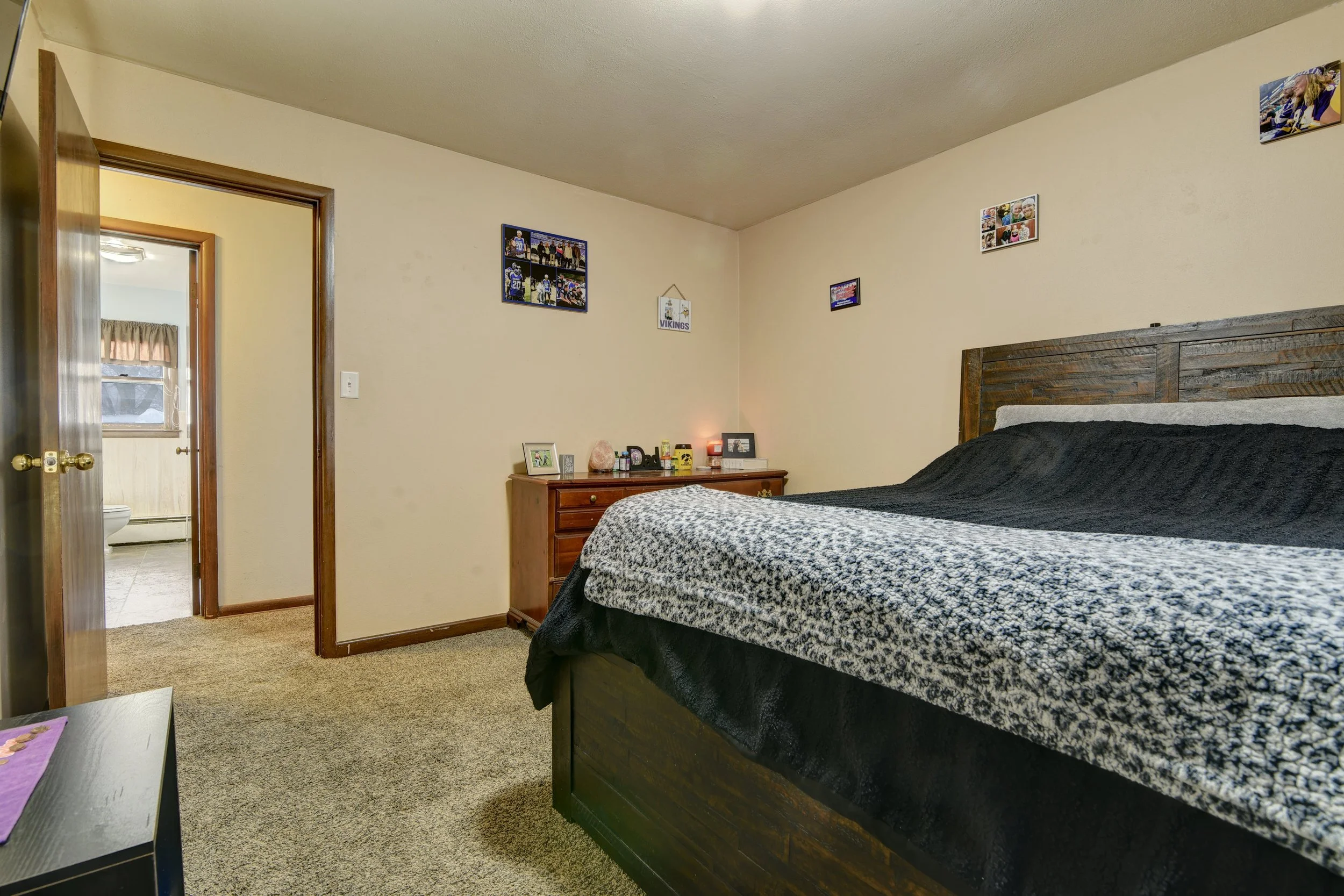 A bedroom with beige walls, a wooden bed frame, and a black and white patterned bedspread. There is a wooden dresser with framed pictures and small decor items on top. The doorway leads to a bathroom with a window and beige tiles.