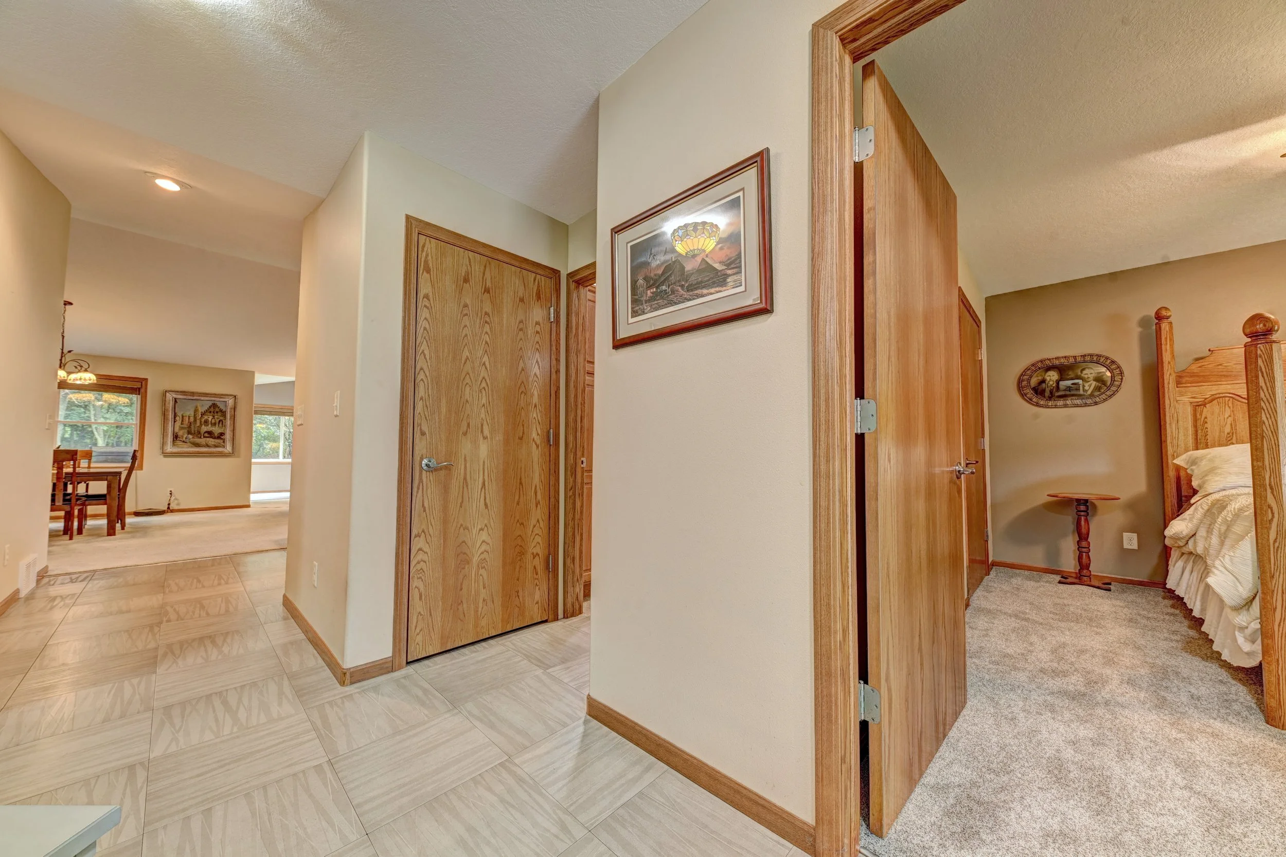 Interior view of a house with a hallway and bedrooms, showing wooden doors, framed pictures on the walls, and a mix of tiled and carpeted flooring.