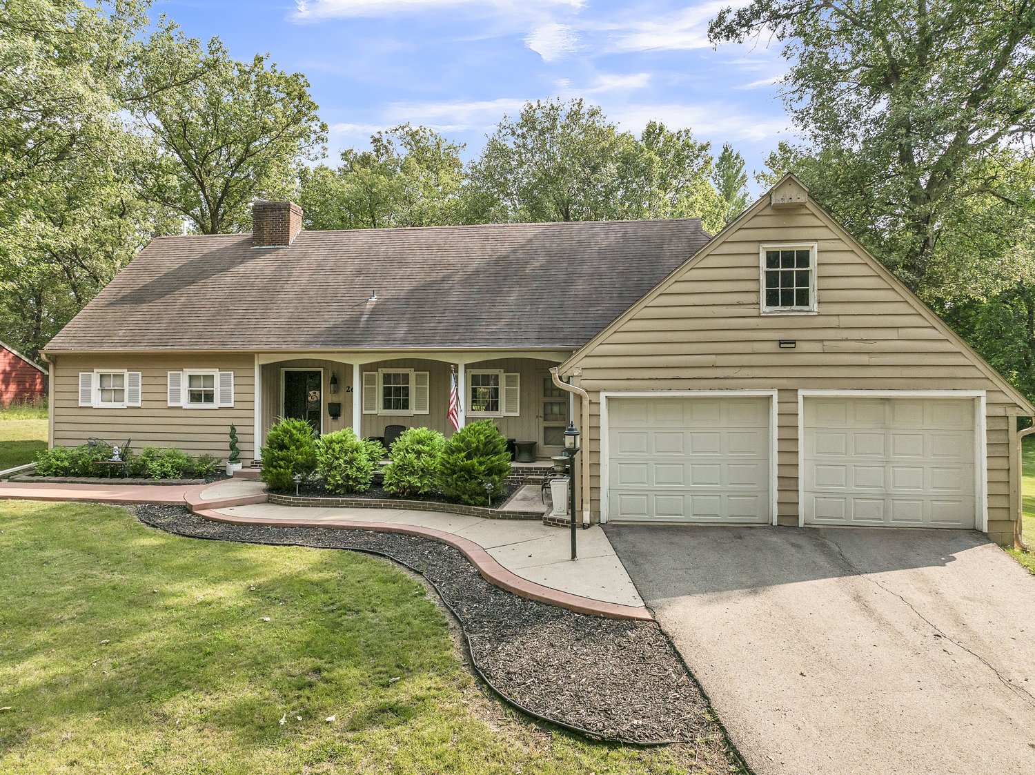 An acreage located in the town of Estherville, Iowa. A beige house with white trim, a two-car garage, a front porch with plants, and a curved walkway. Surrounded by green trees and grass, under a partly cloudy sky.