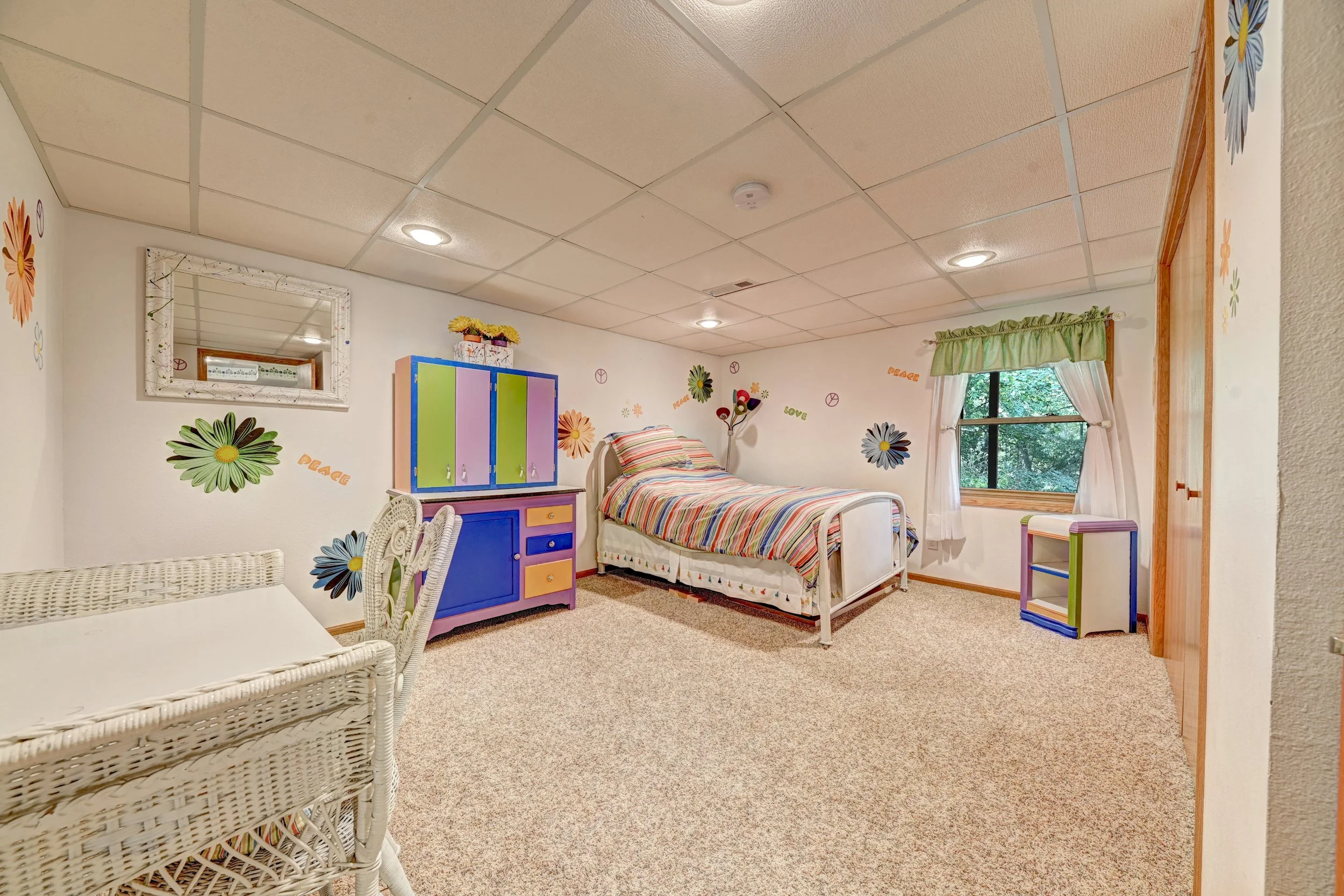 A bedroom with a bed that has a striped multicolored bedspread, decorated wall with colorful flowers and peace signs, a window with white curtains and green valance, a colorful cabinet, and a wicker chair in the foreground.