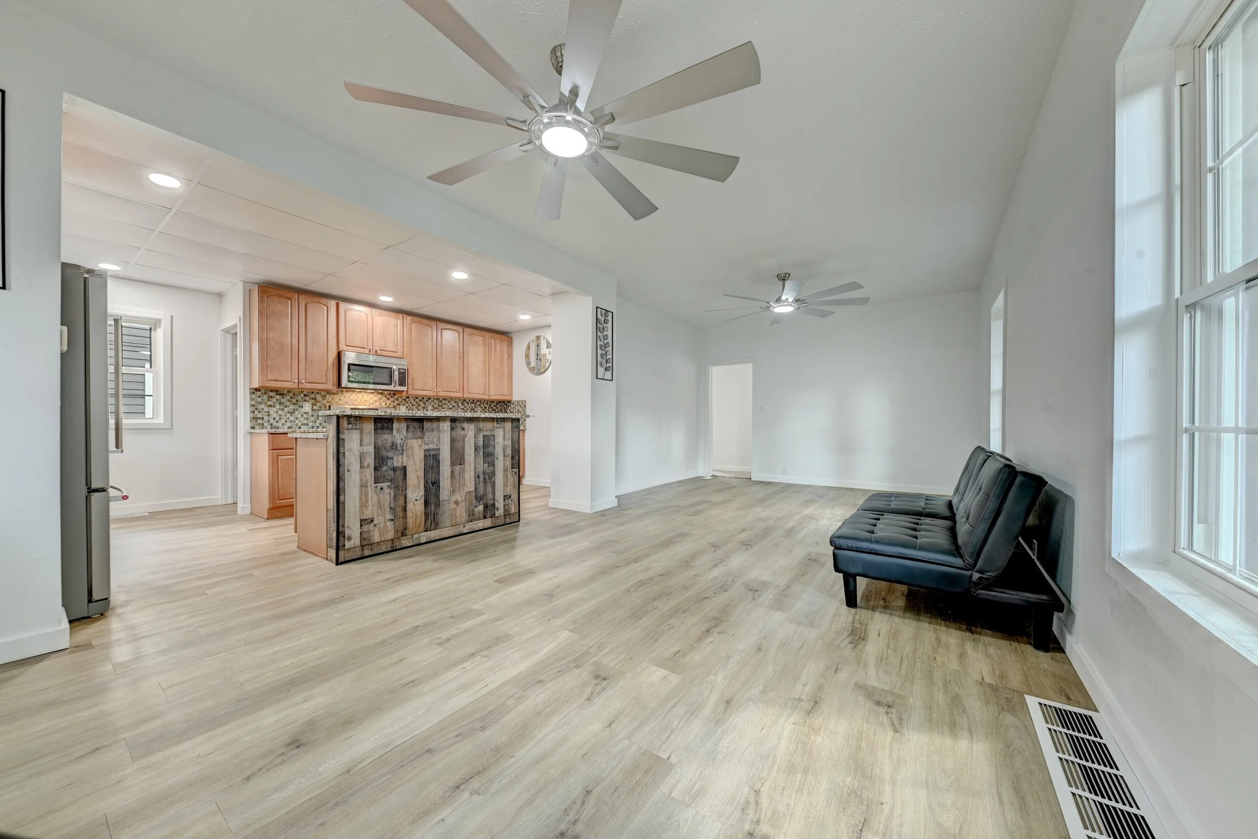 Open-concept living space with a black sofa, light wood flooring, a ceiling fan, and large windows. The kitchen area features wooden cabinets, a microwave, and a rustic island.