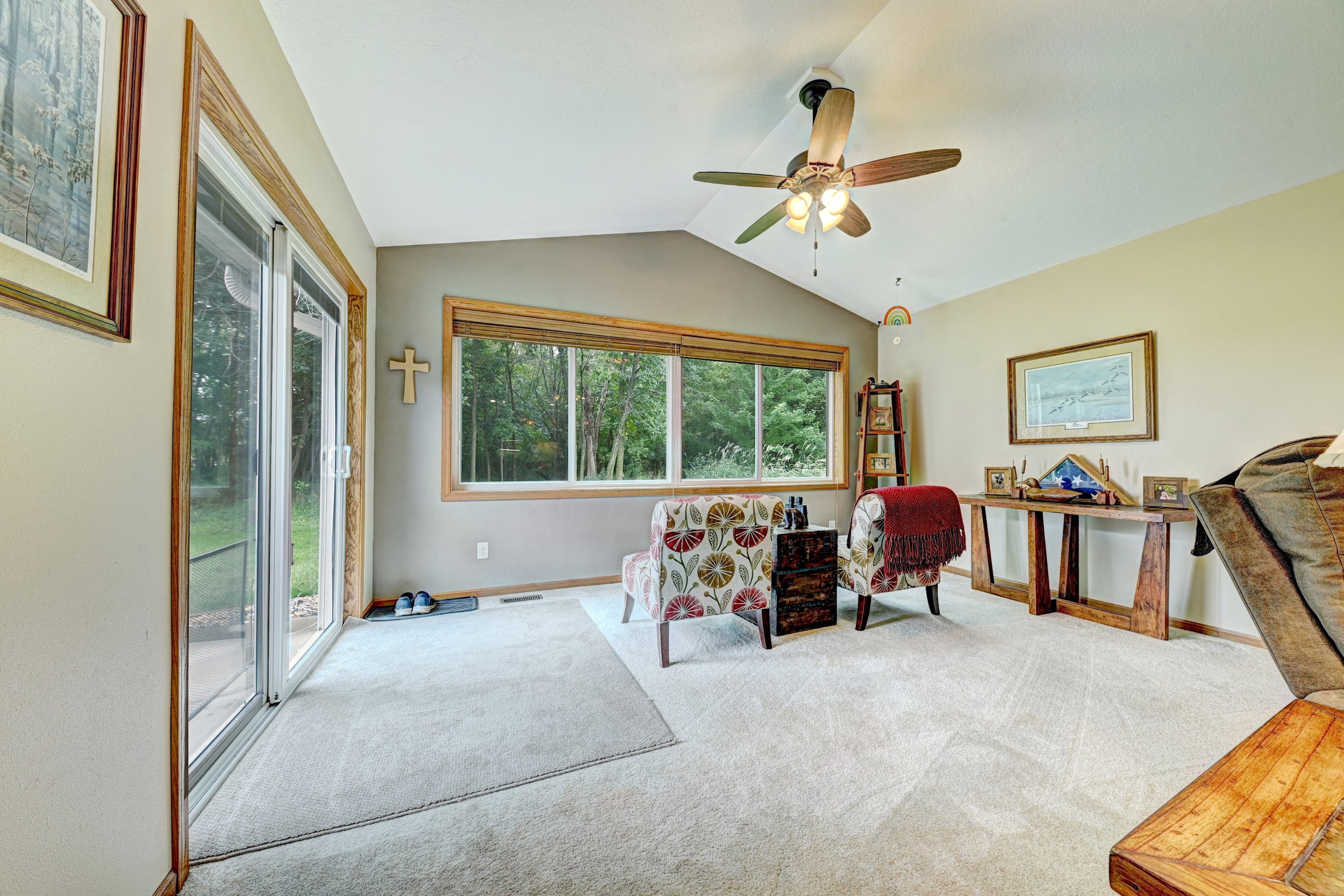 Interior view of a living room with a large window overlooking a green yard, a ceiling fan with lights, a cross on the wall, two patterned armchairs, a wooden shelf with picture frames, and a wooden table with decorative items.