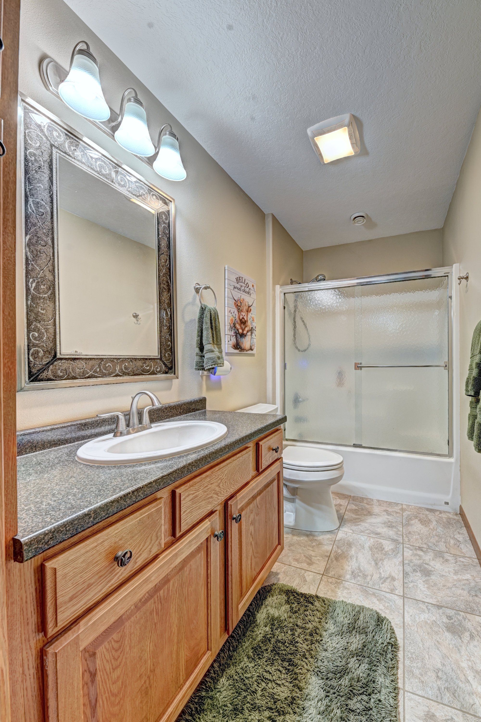 A bathroom with a wooden vanity, a large mirror, a sink, and a toilet. There is a shower with a frosted glass door and a green rug on the tiled floor. The wall has a decorative sign and a towel is hanging on a hook. Overhead lighting is above the mir