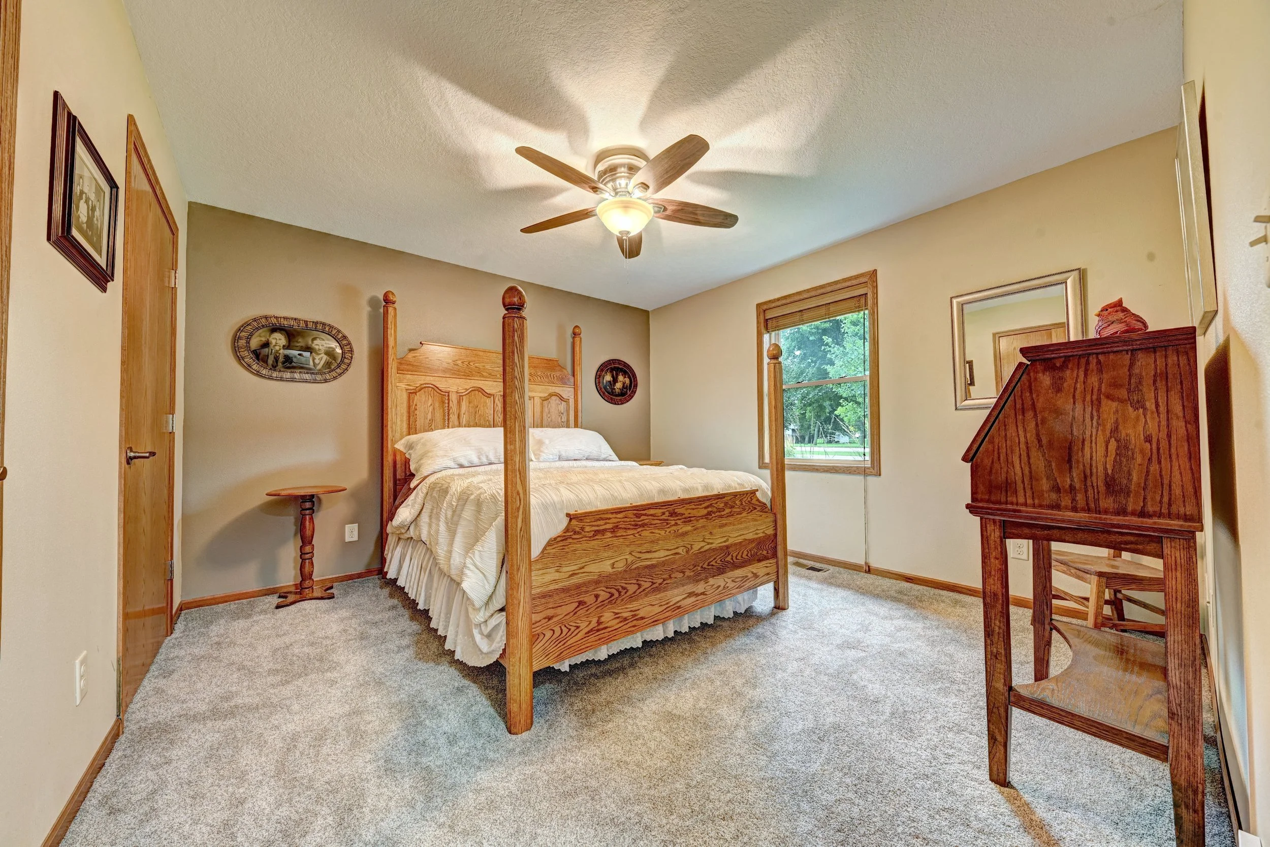 A bedroom with a wooden bed, beige bedding, window with green trees outside, ceiling fan, desks on the side, framed pictures on the walls, and carpeted flooring.