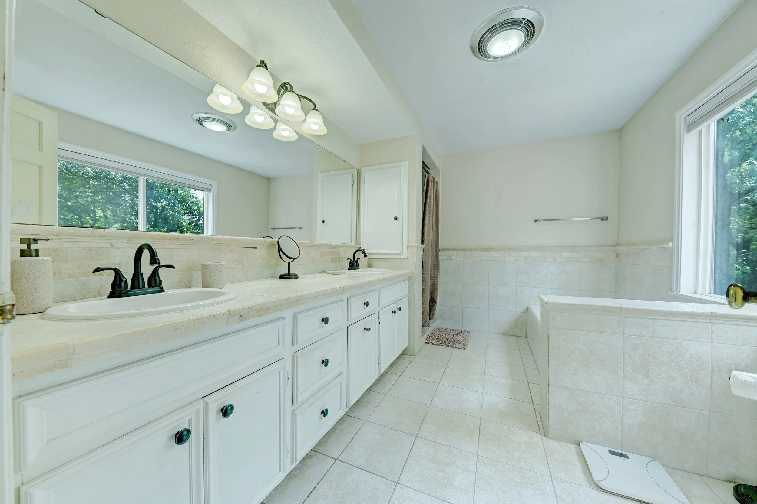Bright bathroom with large mirror, white cabinetry, black fixtures, window with greenery, and a digital scale on tiled floor.