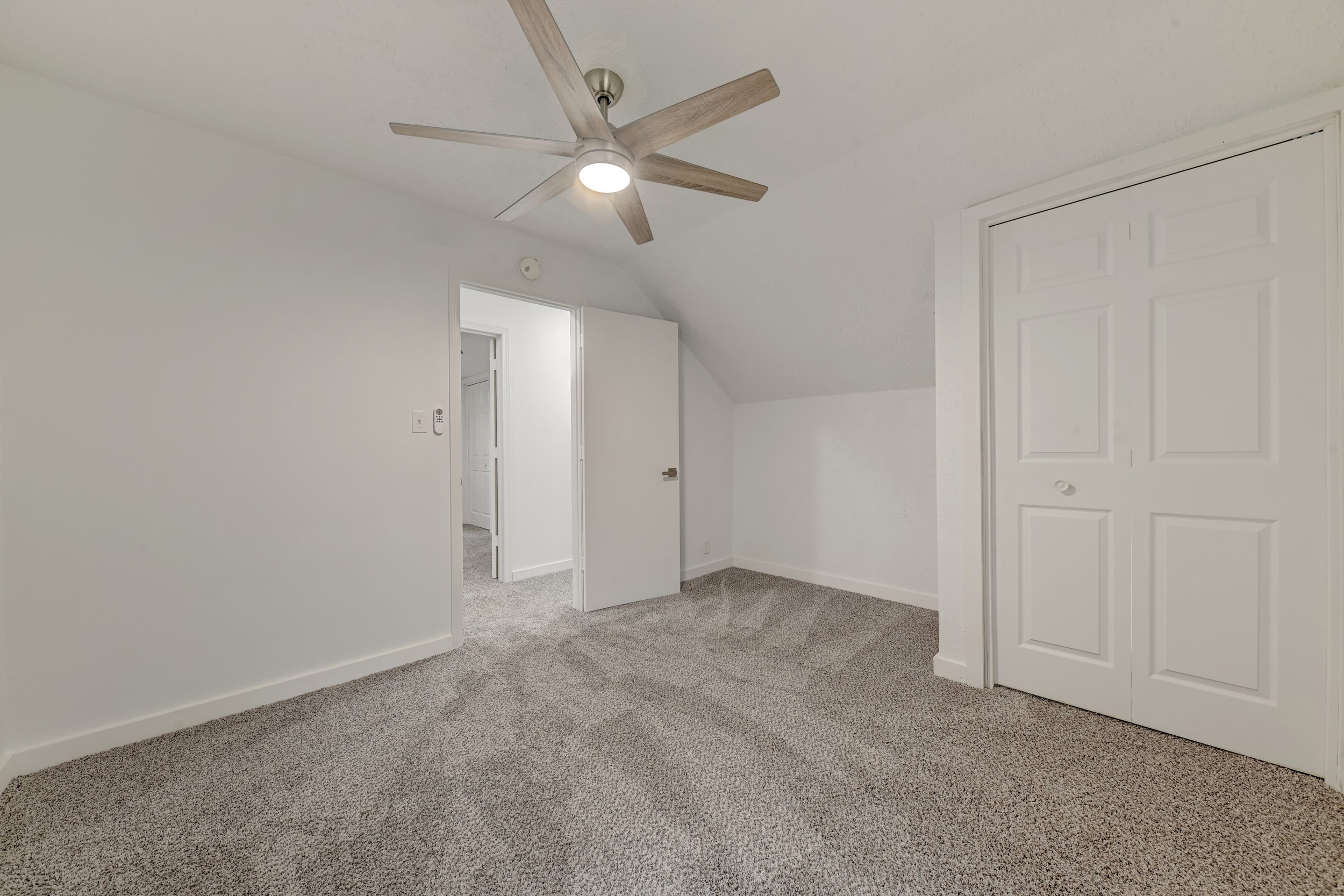 Empty bedroom with white walls, beige carpet, closet doors, ceiling fan, and open door leading to a hallway.