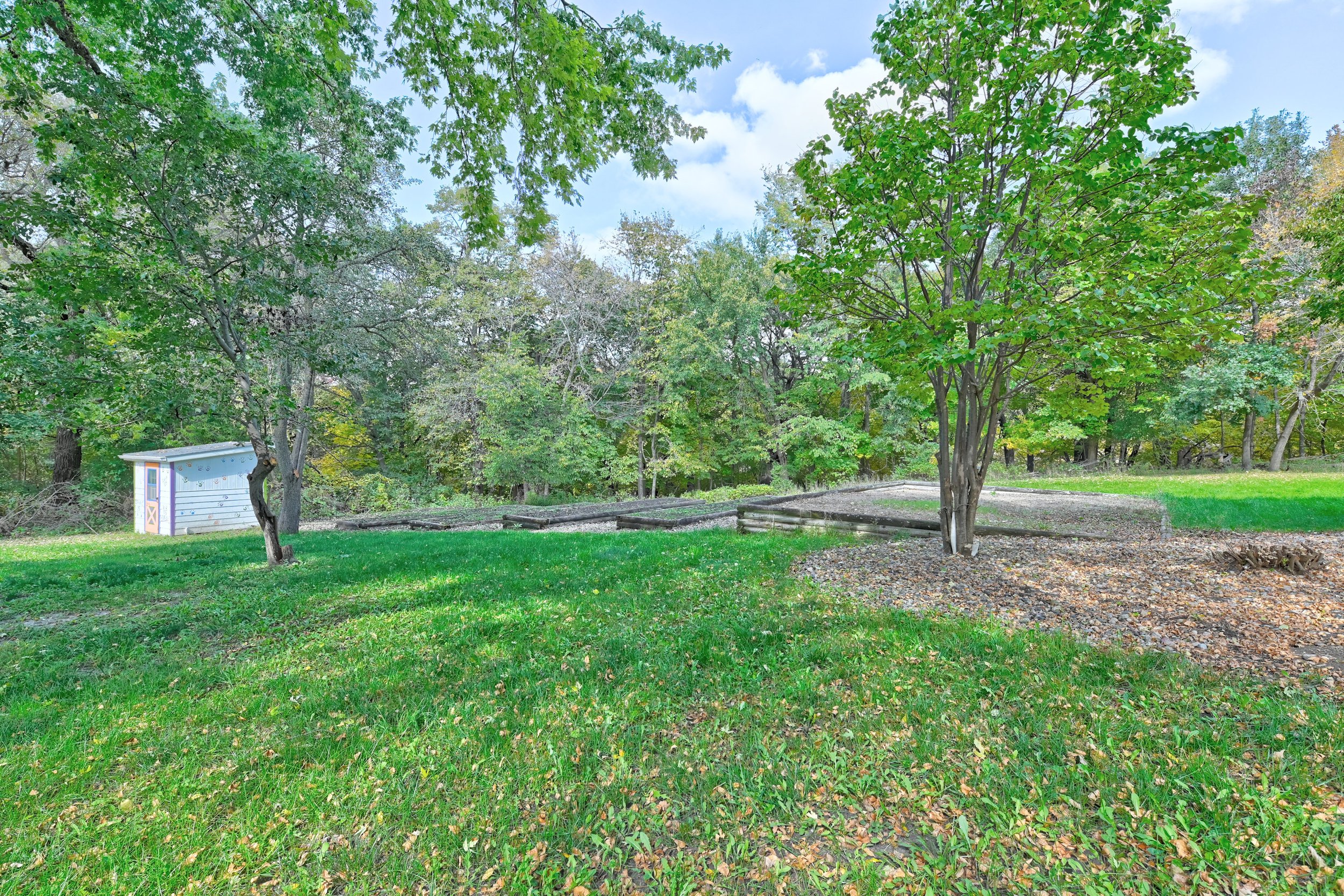 A lush green park with trees, grass, and a small shed in the background, under a partly cloudy sky.