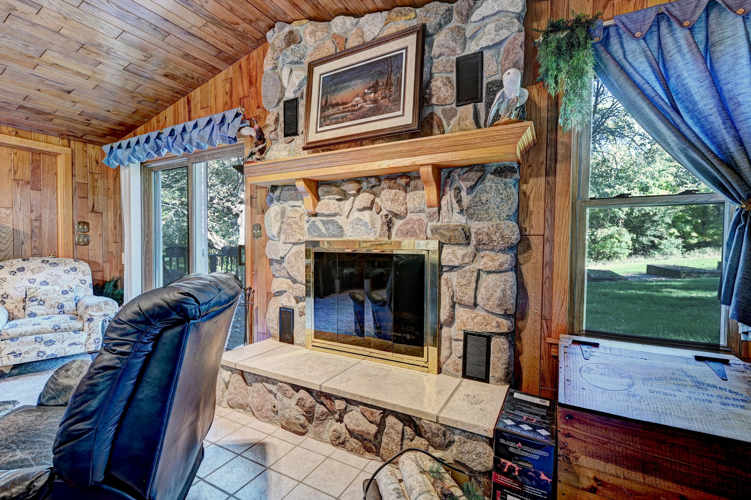 Living room with wood-paneled walls, a stone fireplace with a wooden mantel, and large windows showing a green outdoor scene. Decor includes a framed painting, a decorative bird sculpture, and curtains.