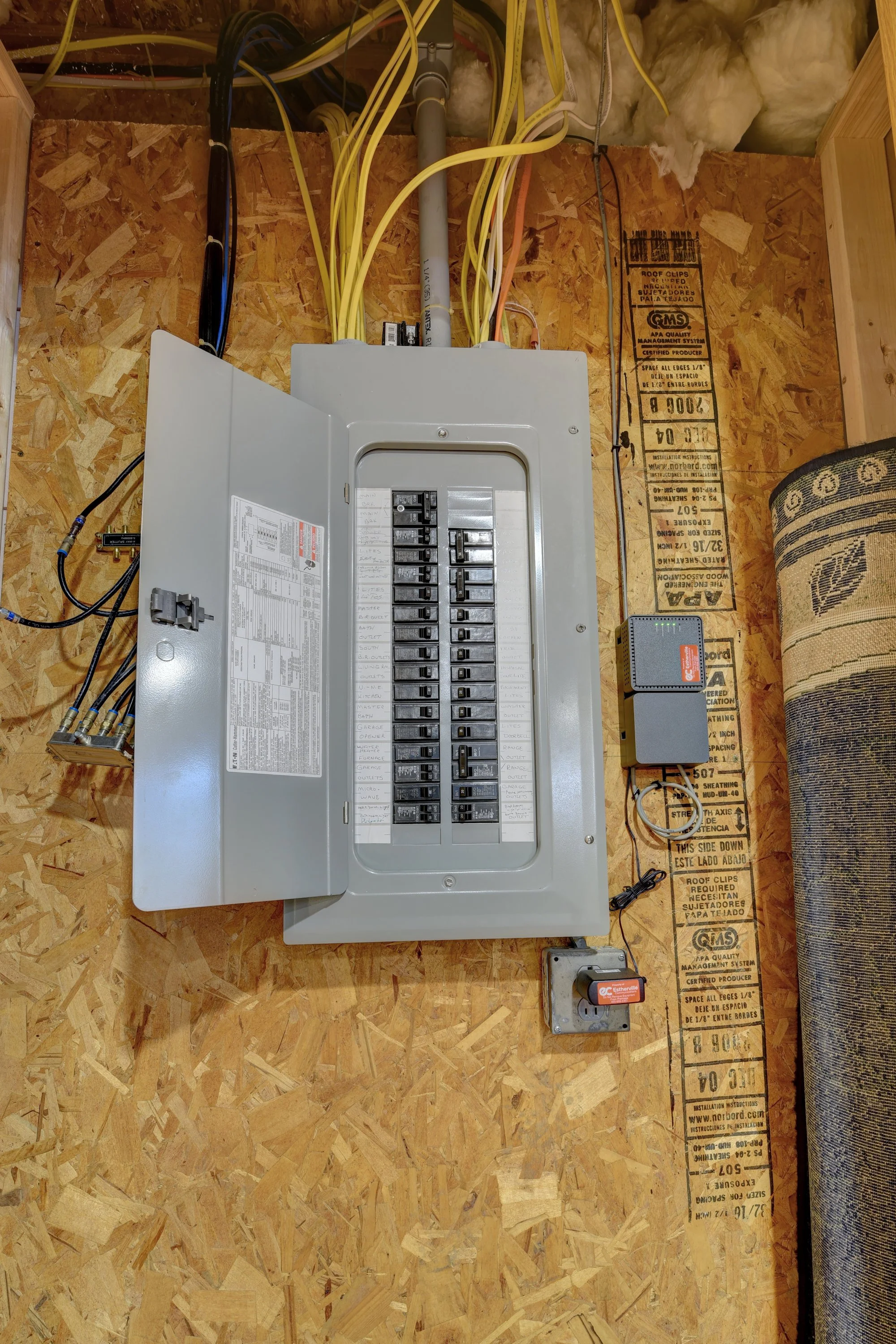 Electrical circuit breaker panel with multiple switches and wires, mounted on a wooden wall next to a rolled-up carpet.