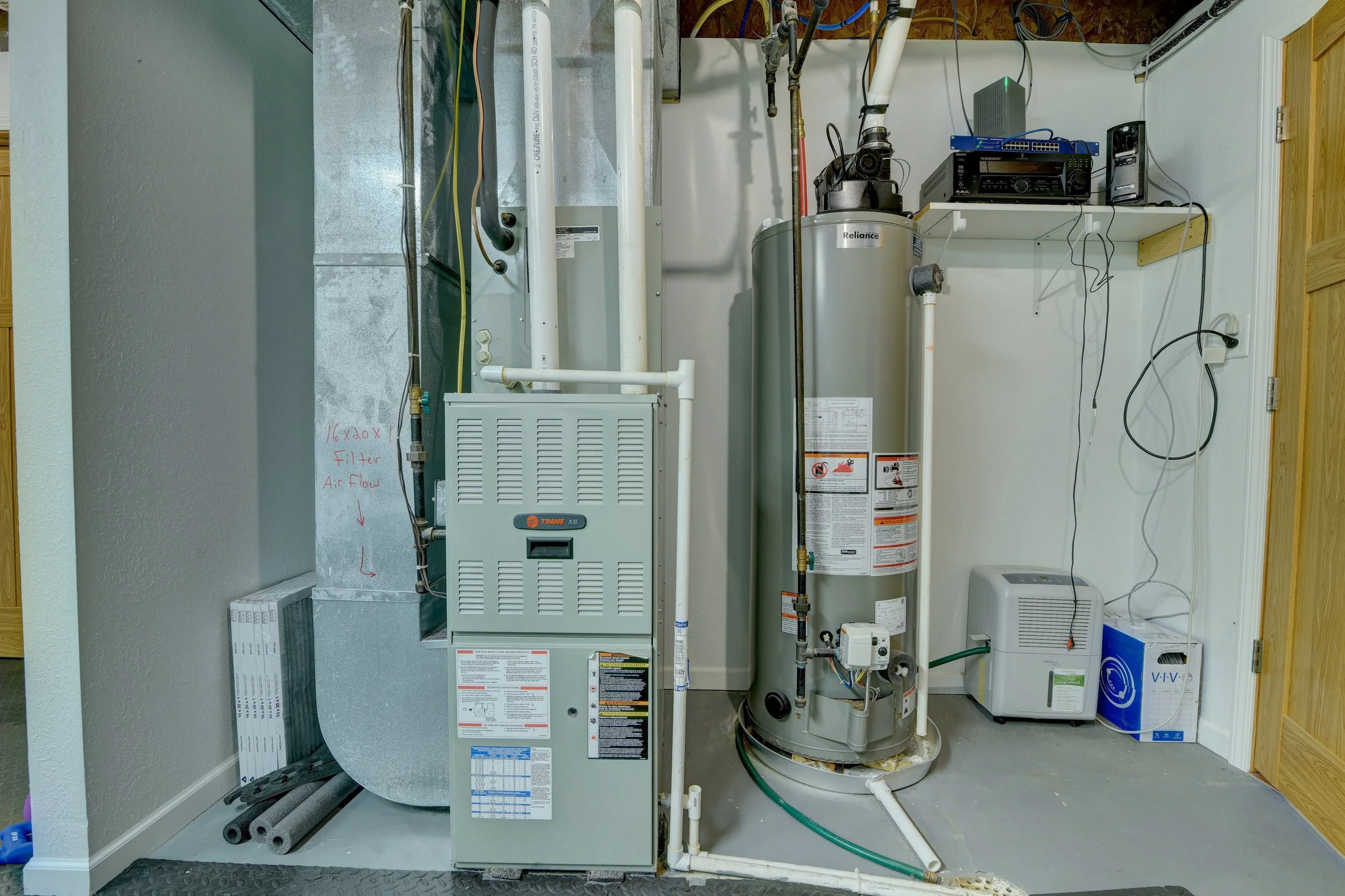 A utility room with HVAC equipment, a hot water heater, and miscellaneous electronic devices, all positioned against a white wall with some pipes and cords.