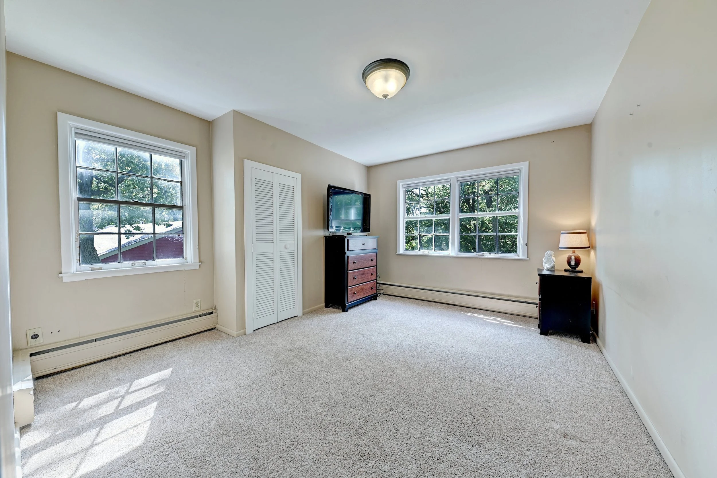 A bright living room with beige walls, two large windows showing trees outside, a ceiling light fixture, a small black dresser with a TV, a lamp, and decorative items on a side table, and white trim and carpet flooring.