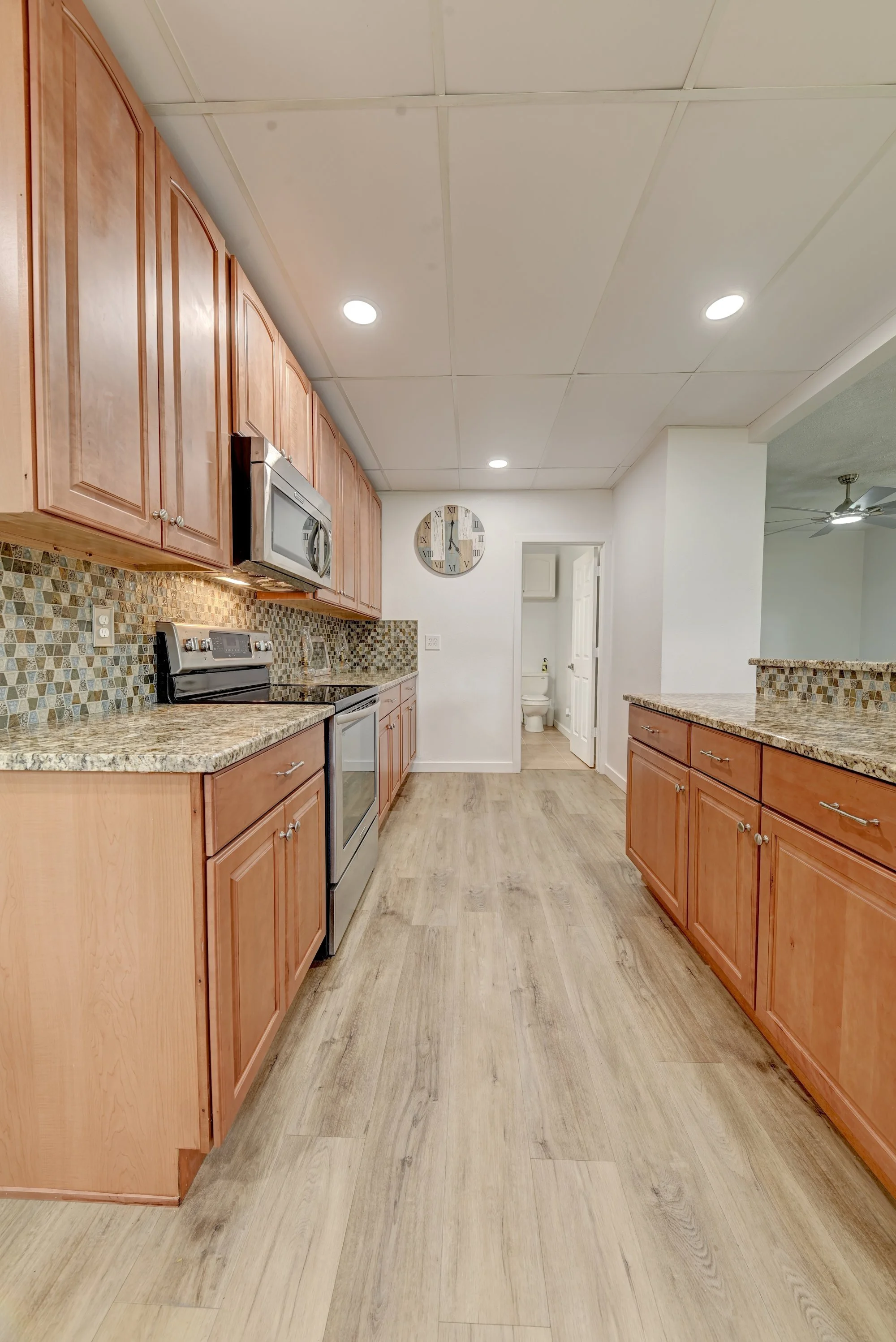 A kitchen with wooden cabinets, granite countertops, stainless steel appliances, mosaic tile backsplash, a clock on the wall, and a view into a bathroom with a toilet.
