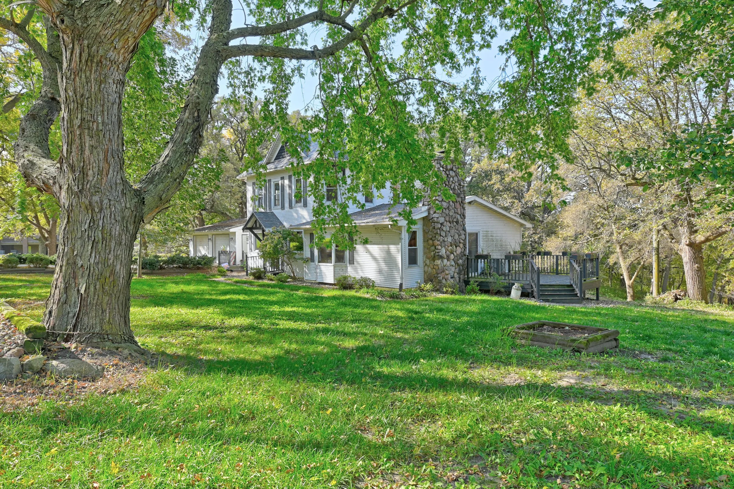 A house with a white exterior and a stone chimney, surrounded by green trees and grass, viewed through the branches of a large tree in the foreground.