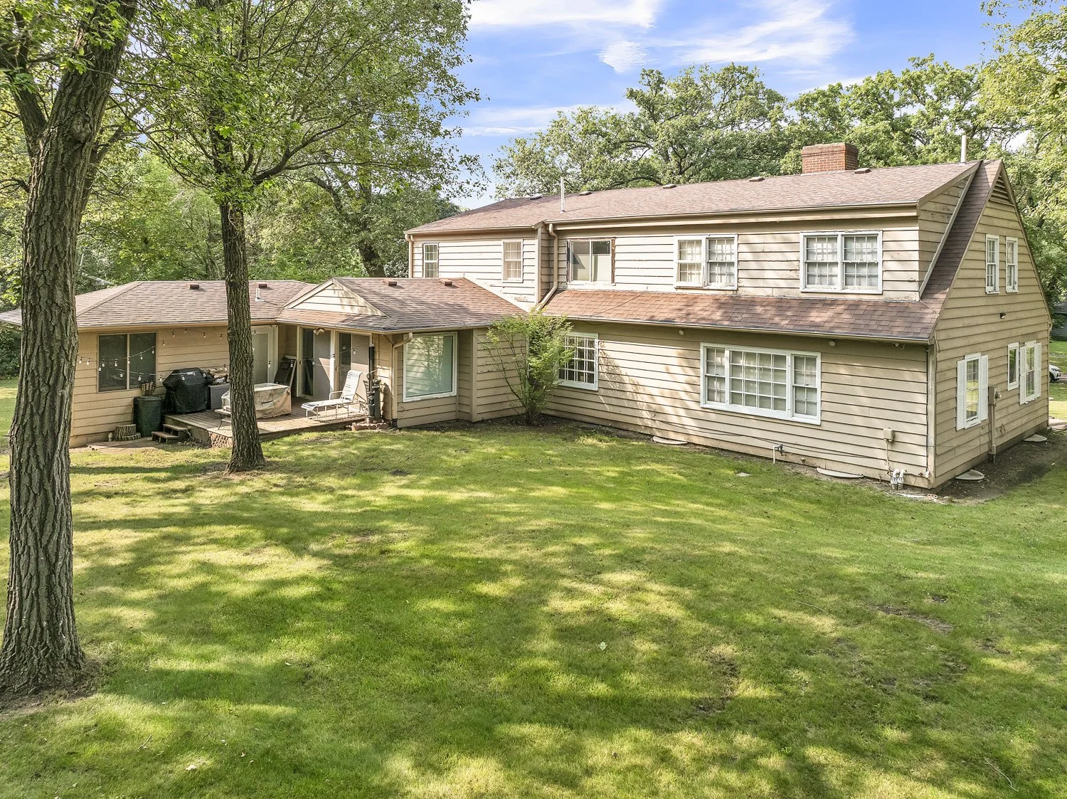 Rear view of a two-story beige house with a small deck, surrounded by green grass and trees, with a blue sky overhead.