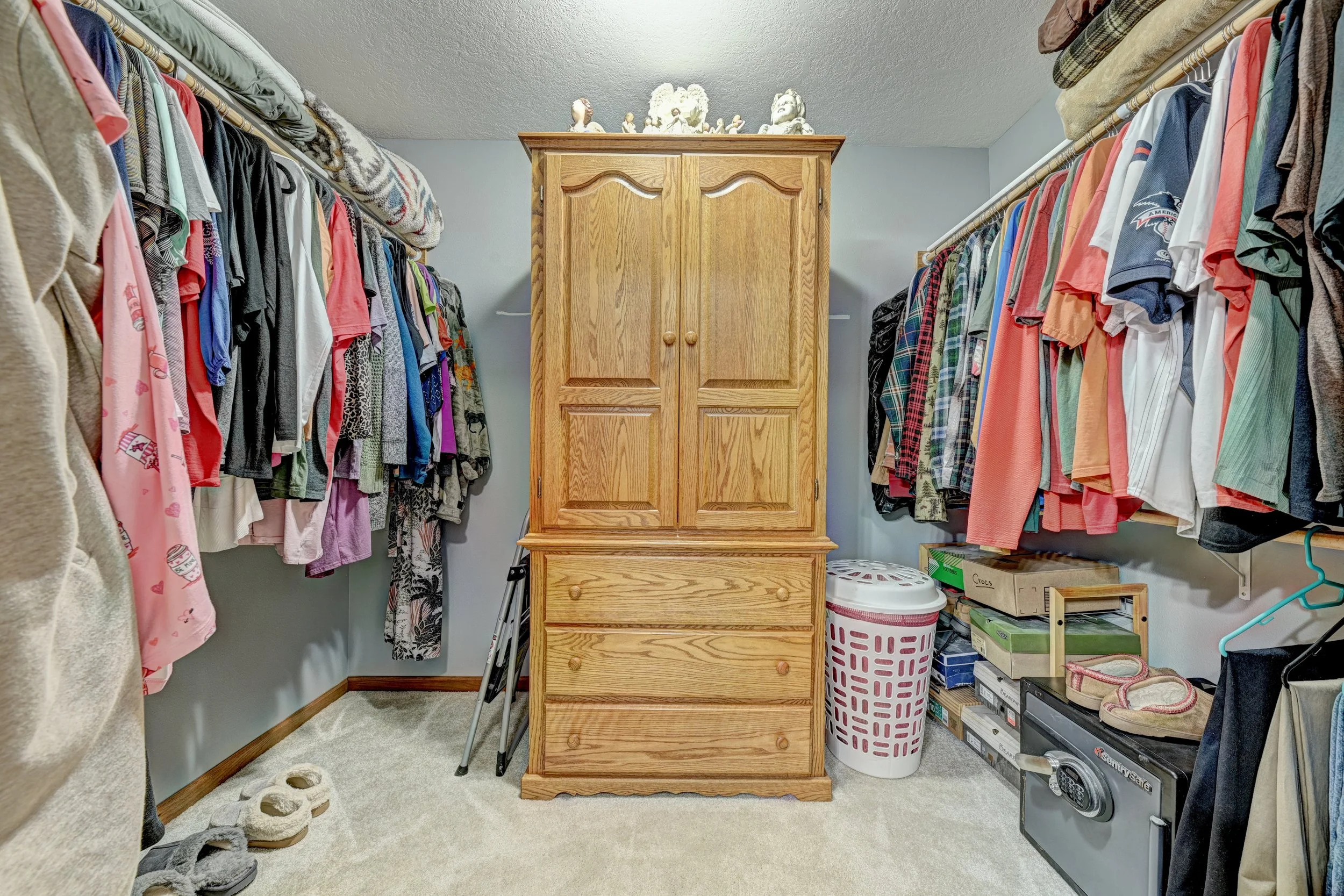 A walk-in closet with two parallel clothing racks filled with various clothing items, a wooden cabinet in the center, and storage boxes on the right side.
