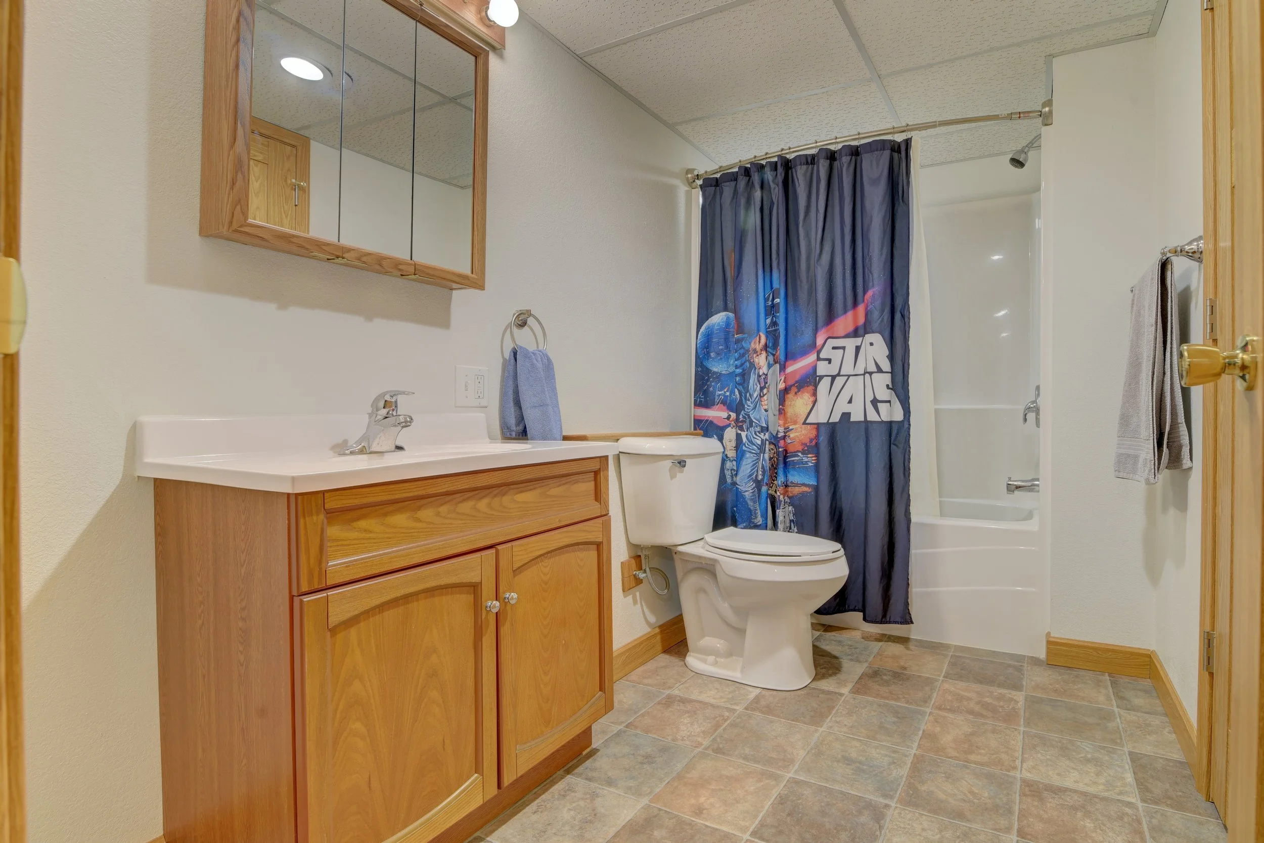 Bathroom with wooden cabinets, a mirror, a white sink, a toilet, a shower with a Star Wars themed shower curtain, and tiled flooring.