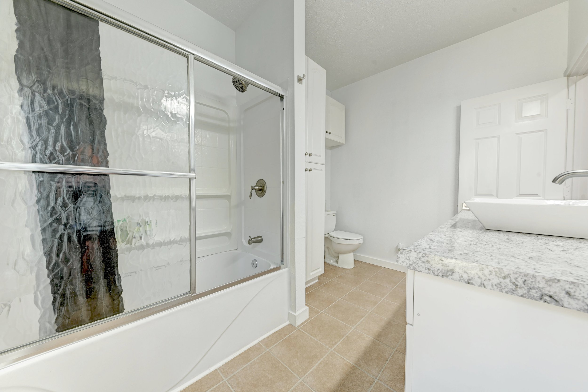 Bathroom with a shower, toilet, white cabinets, granite countertop, and beige tiled floor.
