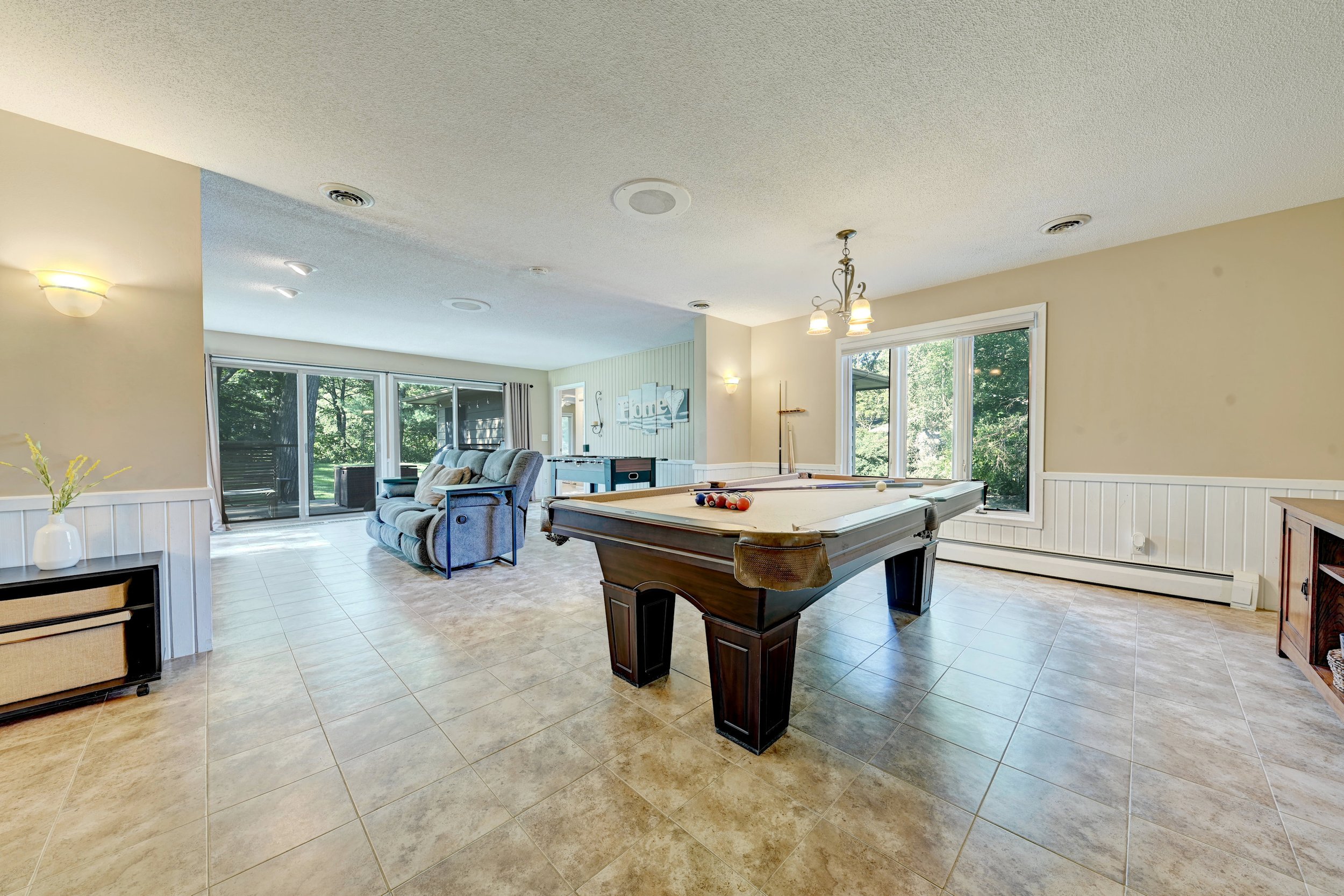 Living room with beige walls, tiled floor, large windows, ceiling lights, pool table, gray sofa, small side table, and a sliding glass door leading outside.