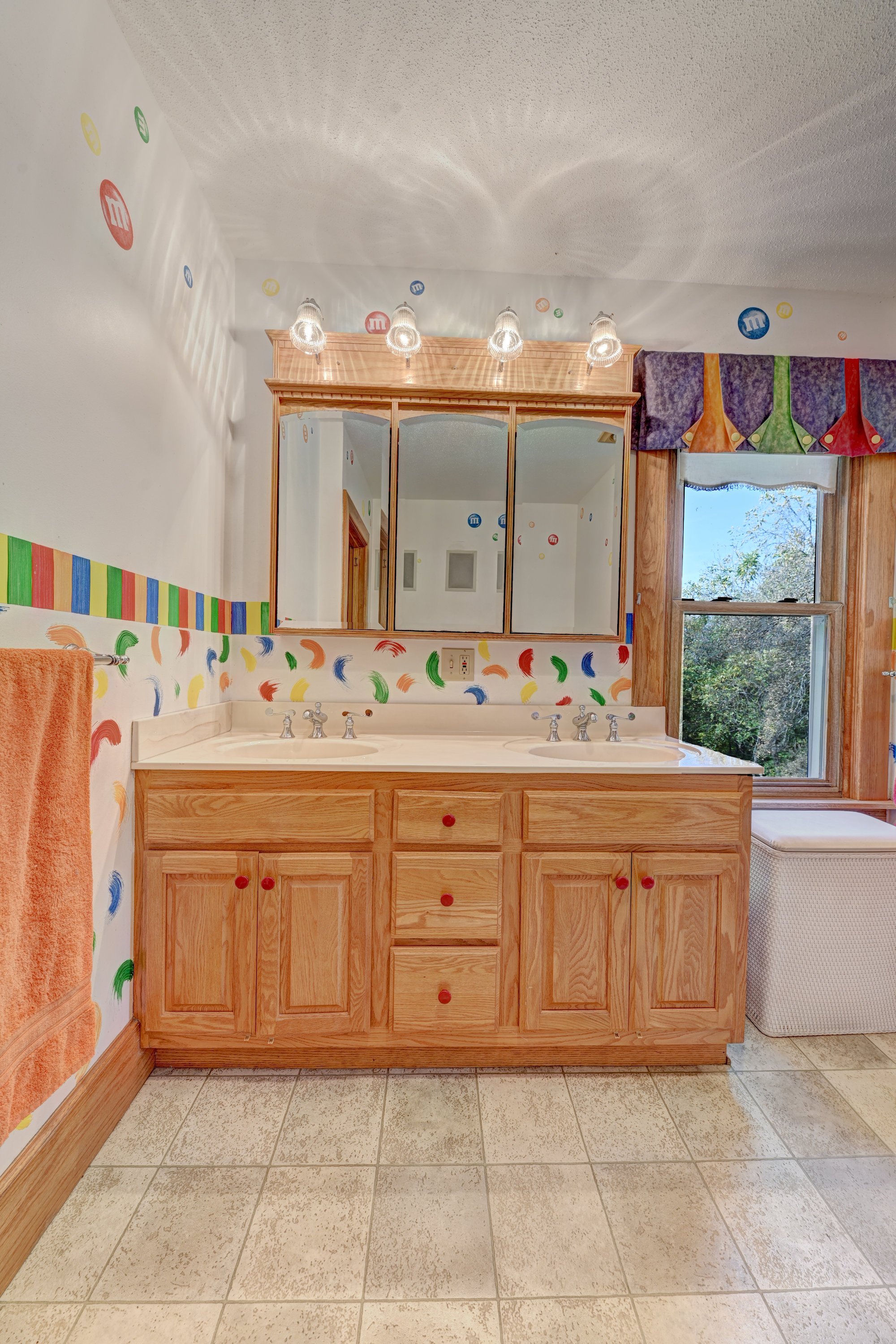 Bathroom with wooden vanity, large mirror, colorful mural, and window showing trees outside.