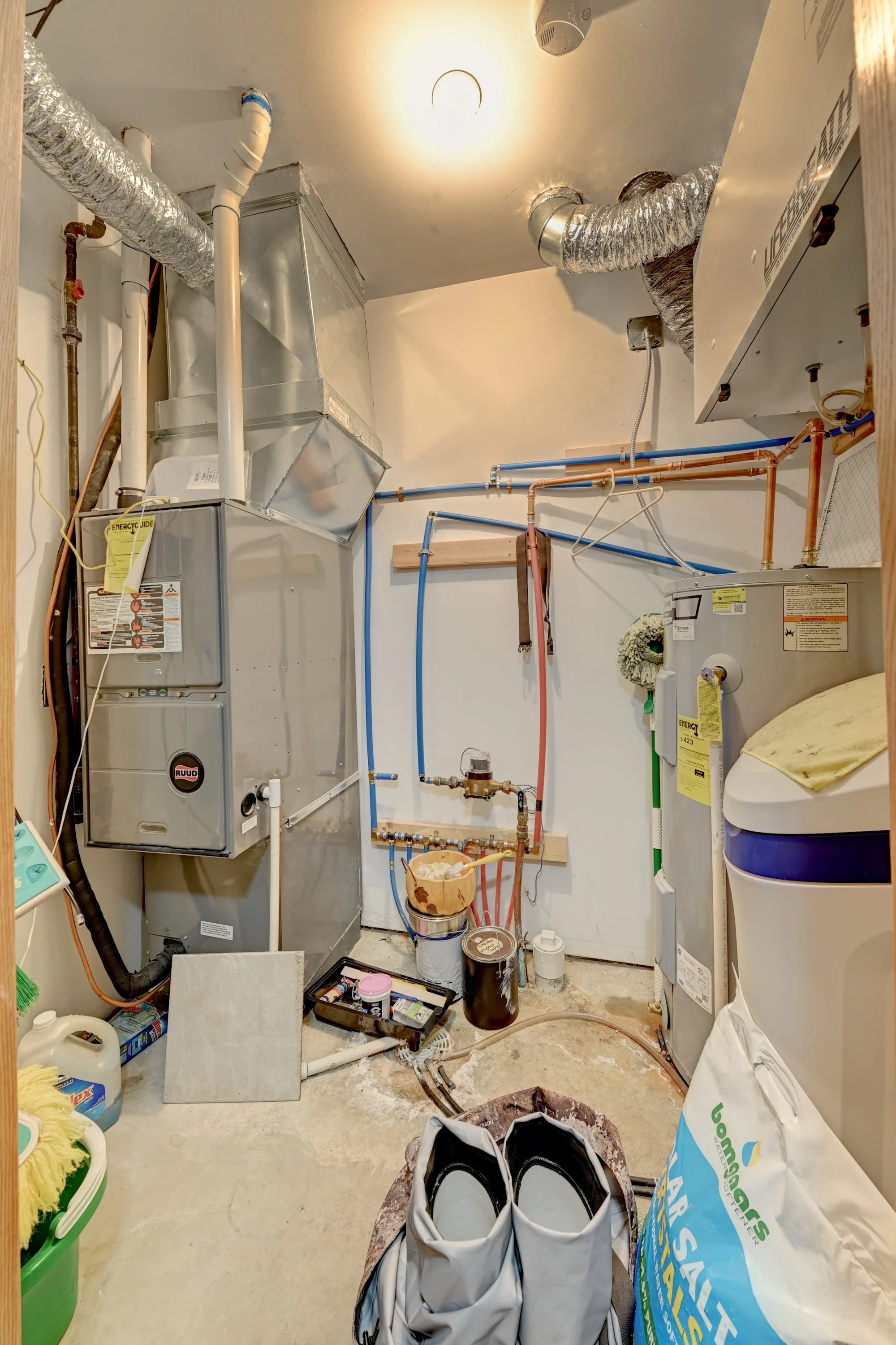 A utility room with HVAC ducts, a water heater, plumbing pipes, cleaning supplies, shoes, and various tools and equipment.