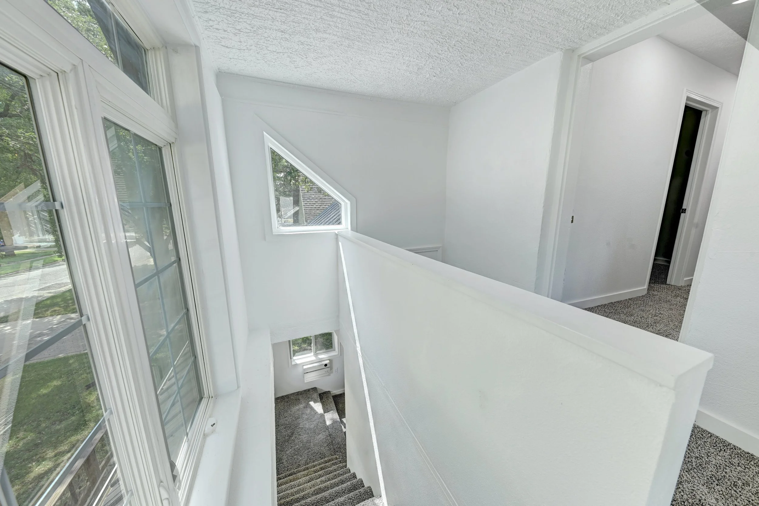Interior view of a white staircase with carpeted steps, leading down from a landing with large windows nearby, and a door on the right side.