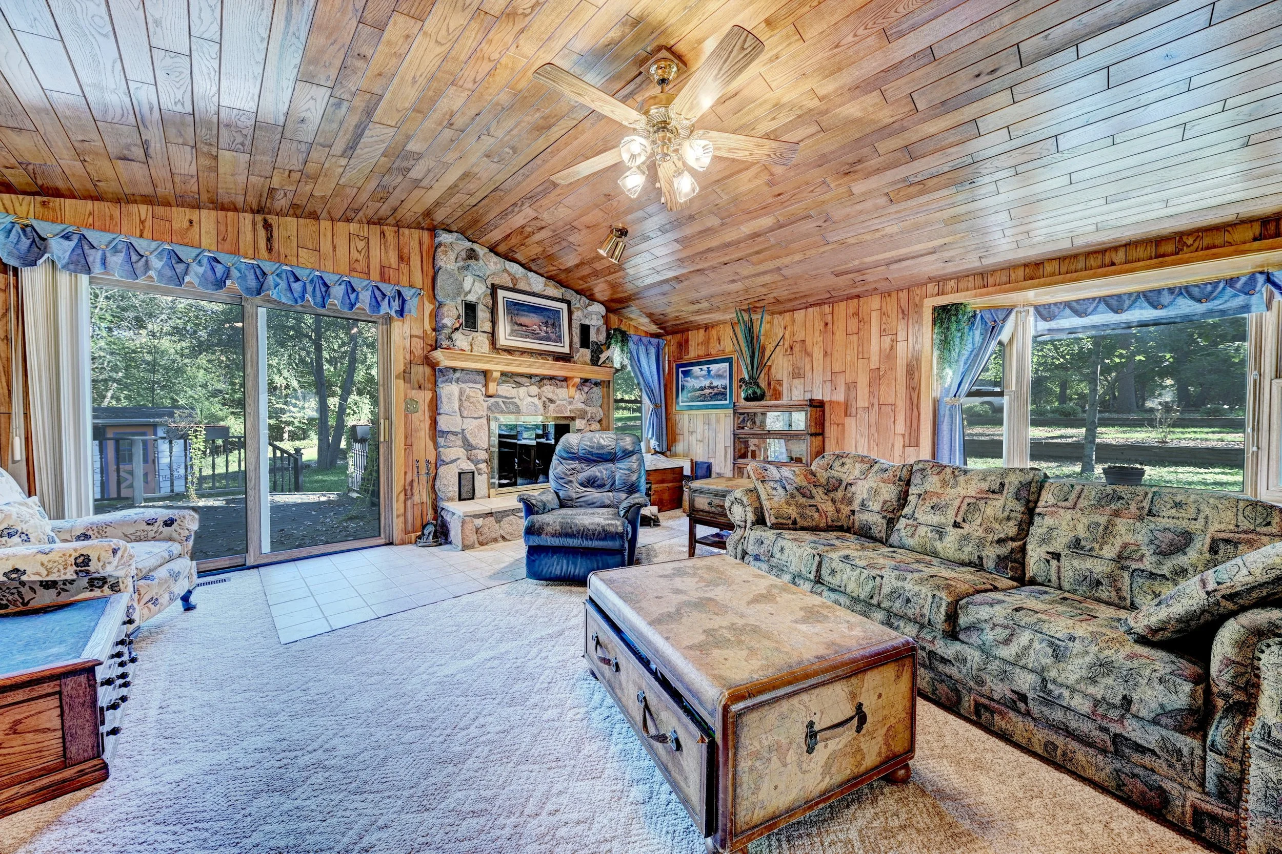 Living room with wood-paneled ceiling and walls, stone fireplace with paintings above, large windows with blue curtains showing a green outdoor scene, floral patterned couches, a leather armchair, a wooden coffee table, a decorative chest, and variou