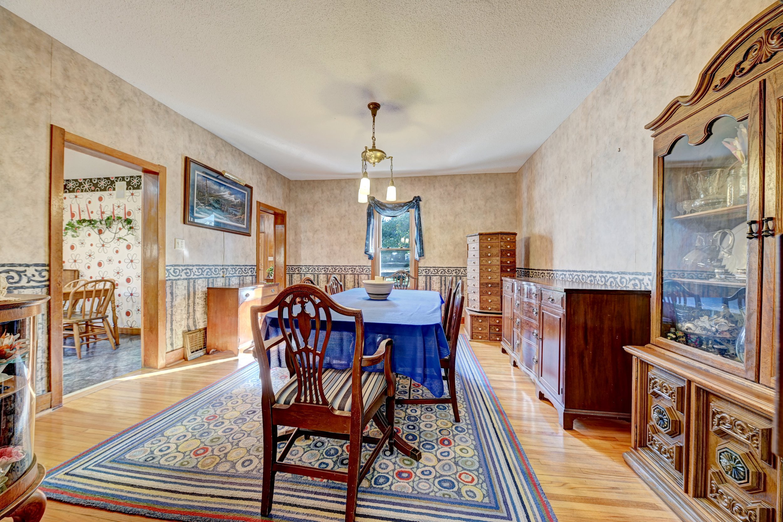 A dining room with a wooden table covered by a blue tablecloth and surrounded by wooden chairs, a hutch on the right, a cabinet on the left, a window with a valance, and a ceiling light fixture.