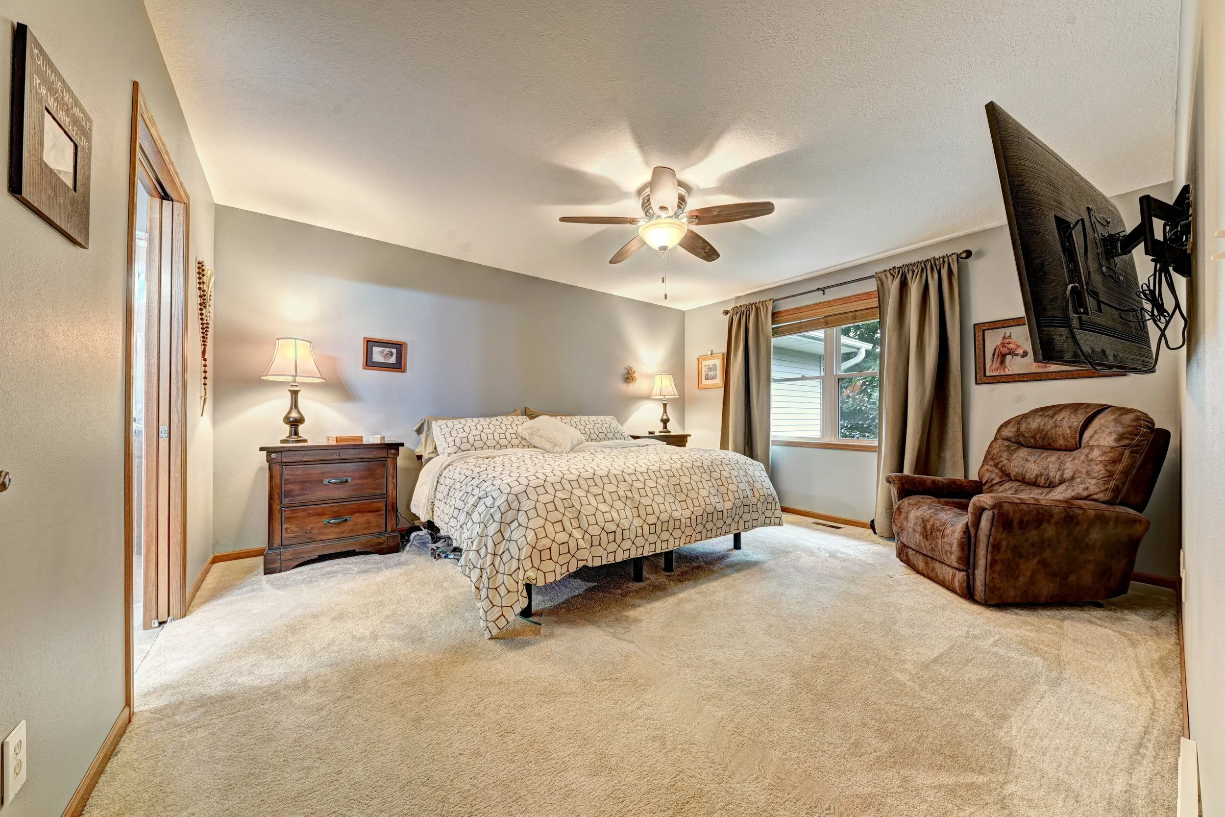 A cozy bedroom with a bed, nightstands, lamps, a wall-mounted TV, and a window with curtains.
