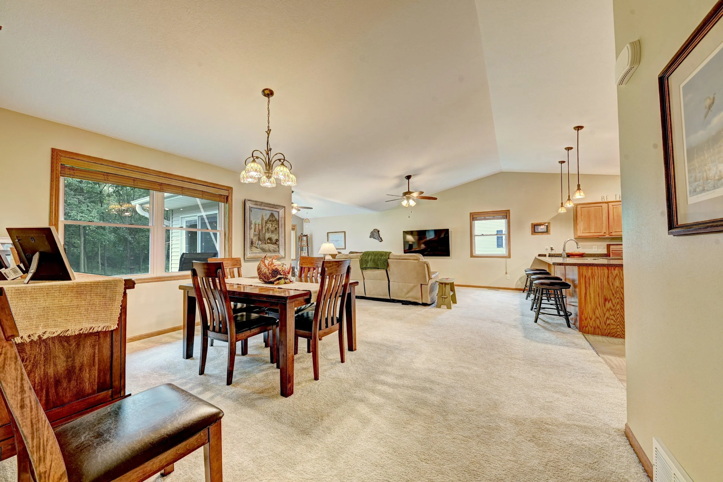 Living room with dining table, chairs, beige carpet, wall art, and kitchen counter, with windows looking to outside greenery.