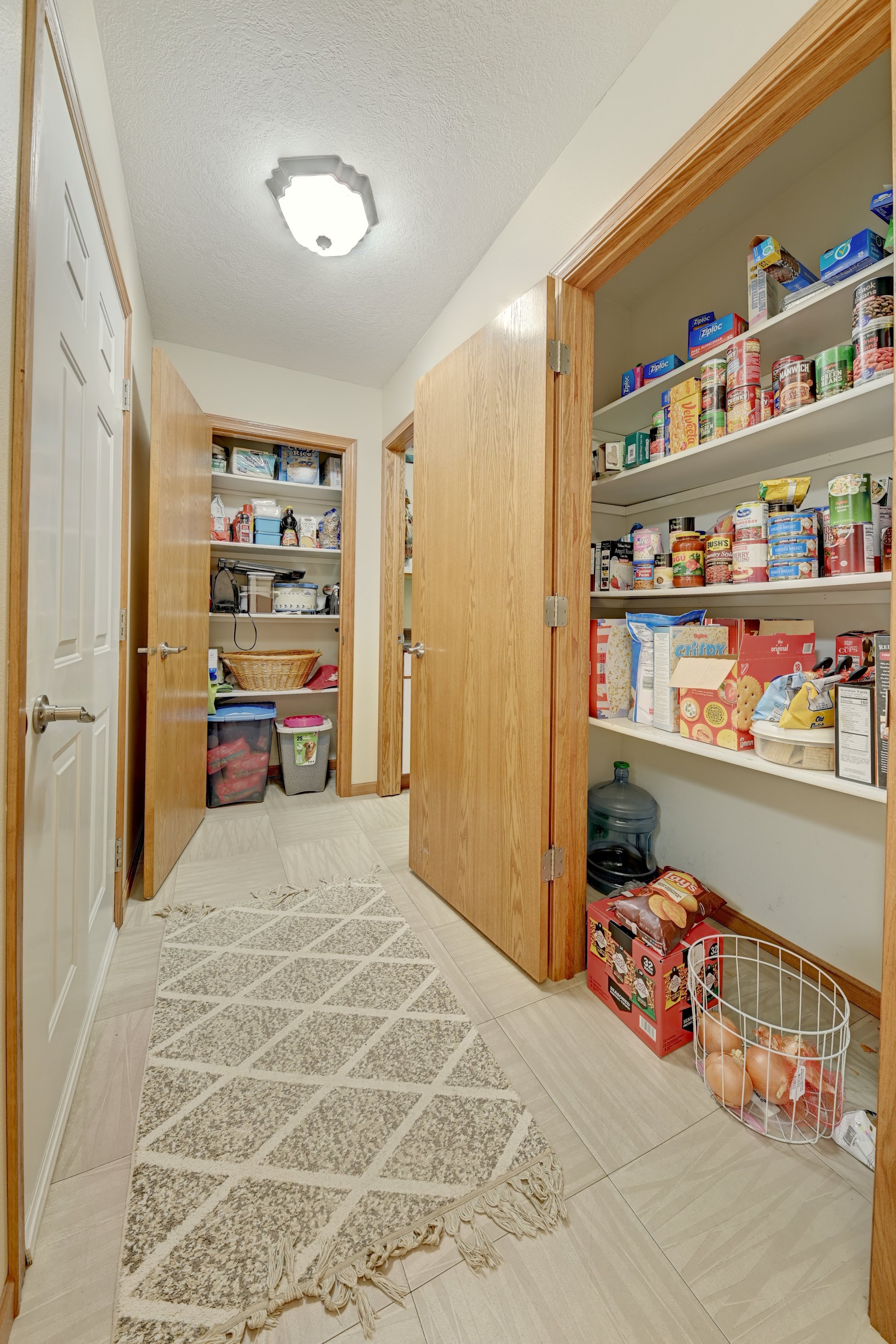 A view inside a pantry closet with open wooden doors, white shelves stocked with canned goods, boxes, and bags of food, and a water jug on the floor. There is a beige patterned rug on the tile floor.