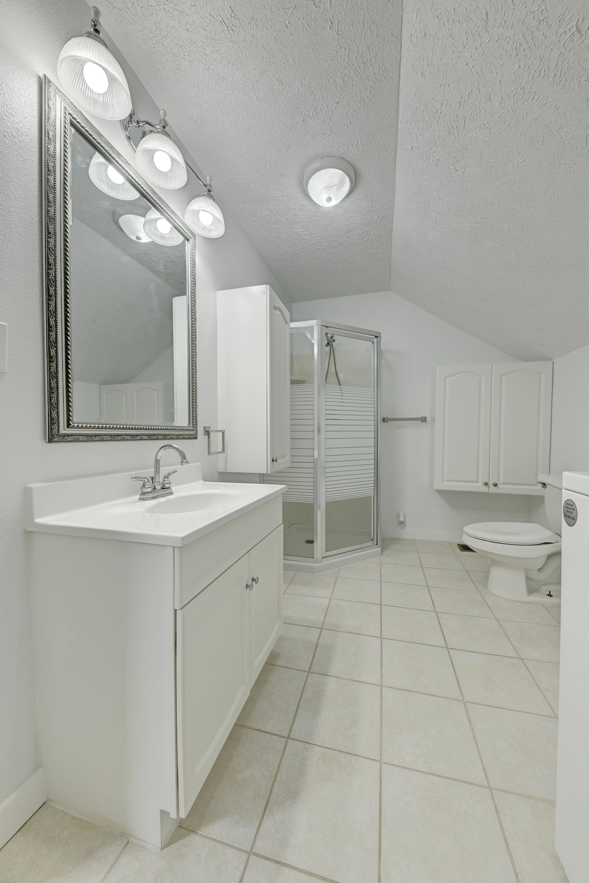 A clean, white bathroom with a vanity, mirror, and lighting fixture, a shower with a glass door, and a toilet, all with beige tile flooring.
