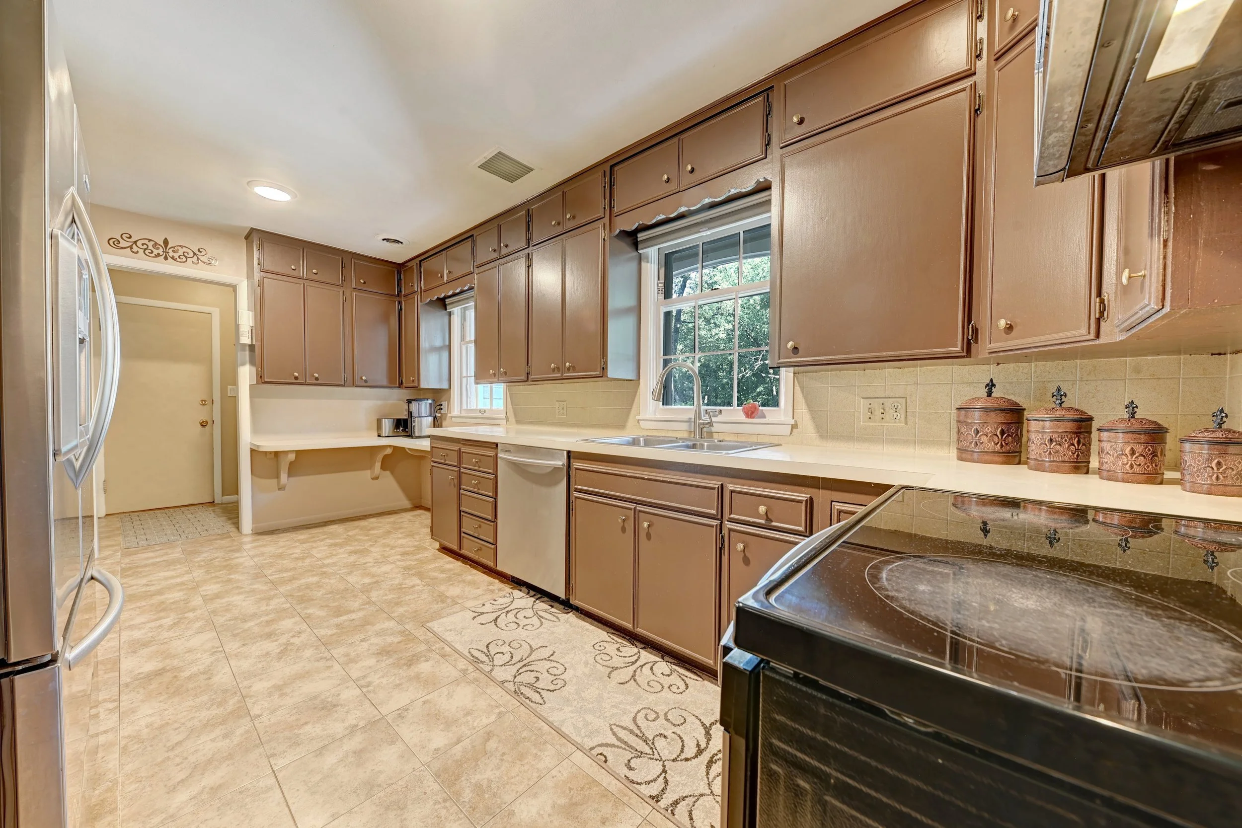 A kitchen with brown cabinets, white countertops, a stainless steel refrigerator, a window above the sink, and decorative jars on the counter.