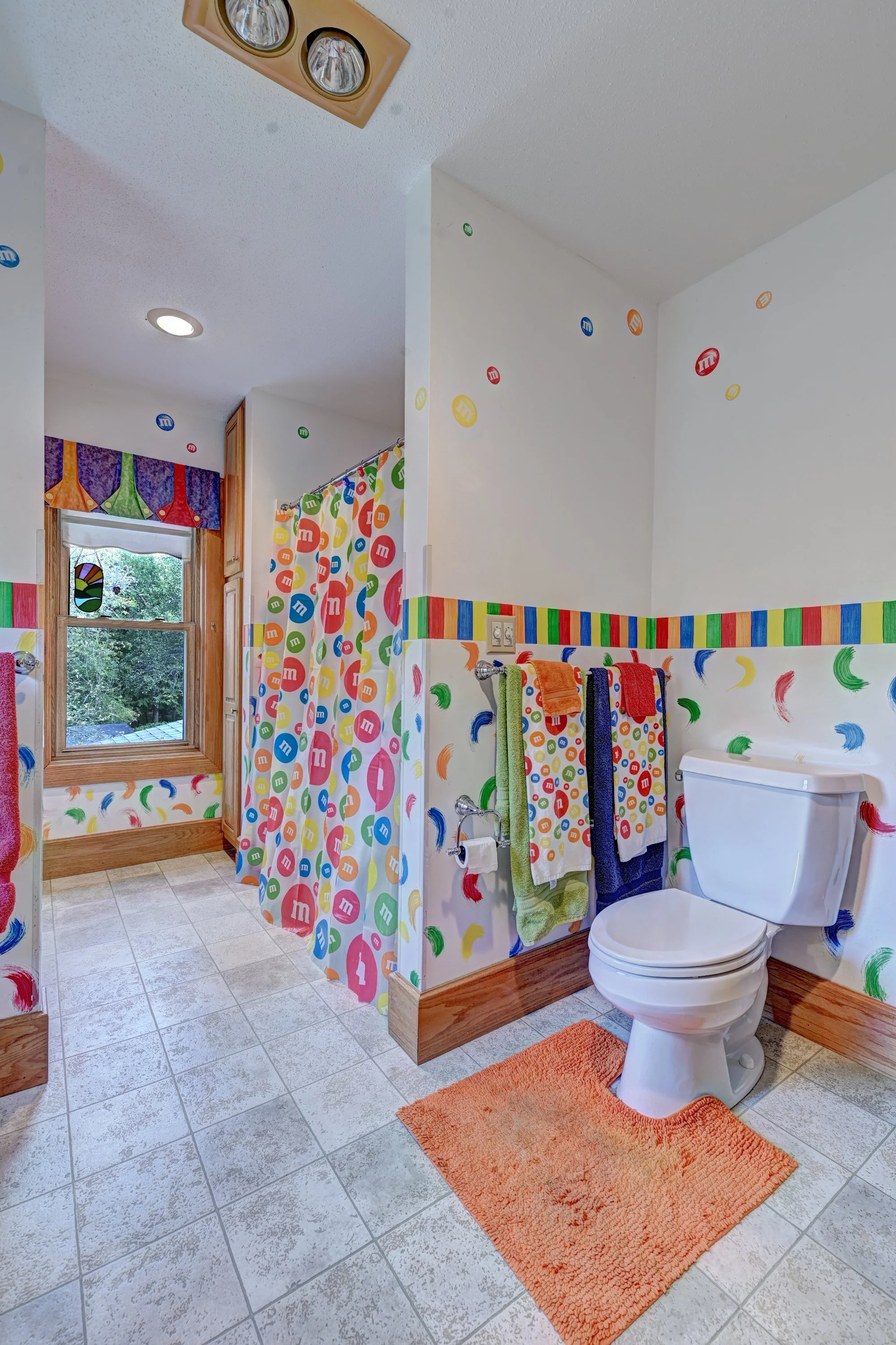 A bathroom decorated with colorful M&M's themed decor. Features a white toilet, orange rug, and multicolored towels hanging on a rack. The walls are adorned with M&M's stickers, and the shower curtain and window curtain are patterned with the candy l