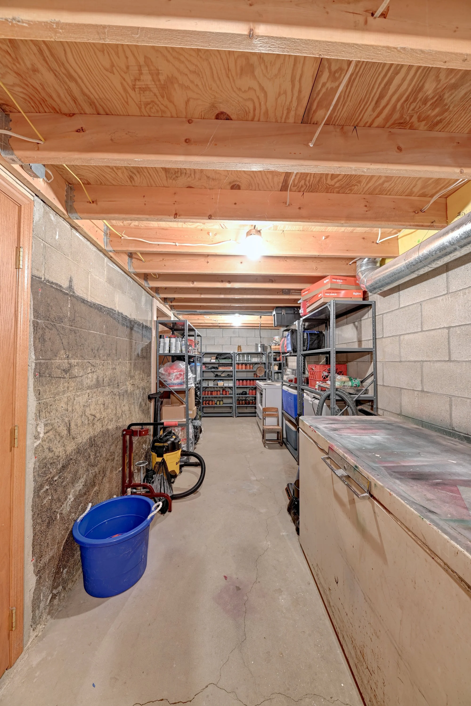 A view of a basement workshop or storage area with a concrete floor, cinder block walls, and a wooden ceiling. Shelving units hold tools, supplies, and boxes. A blue bucket and a yellow wet/dry vacuum are on the floor, and bright lighting illuminates