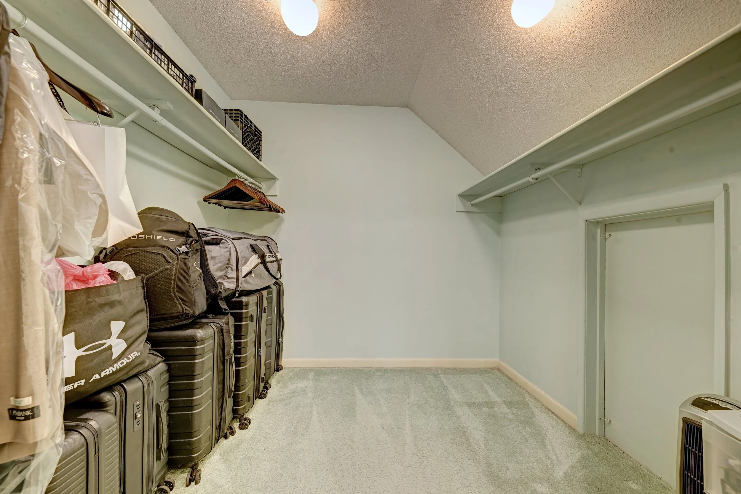 A walk-in closet with luggage, backpacks, and bags stacked on the left. White shelves with black storage bins and coat hangers above. Light green walls, white trim, and beige carpet. Ceiling lights and an air conditioning unit.
