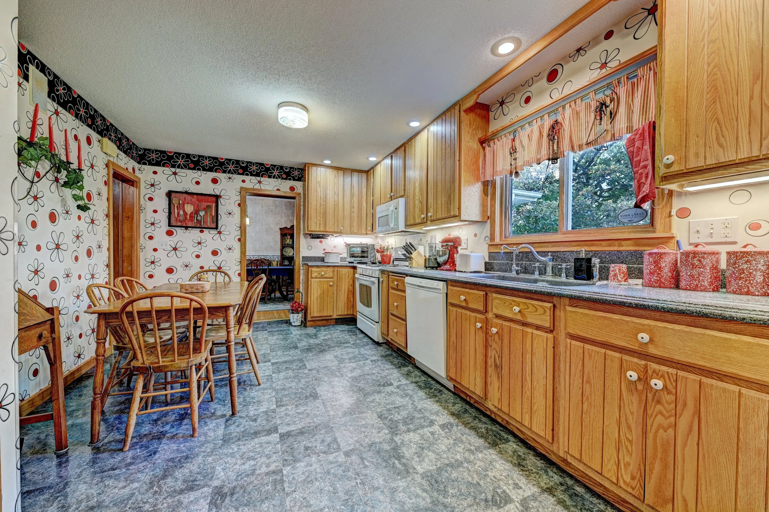 Kitchen with wooden cabinets, a window with floral curtains, a dining table with wooden chairs, and various appliances and decorations.