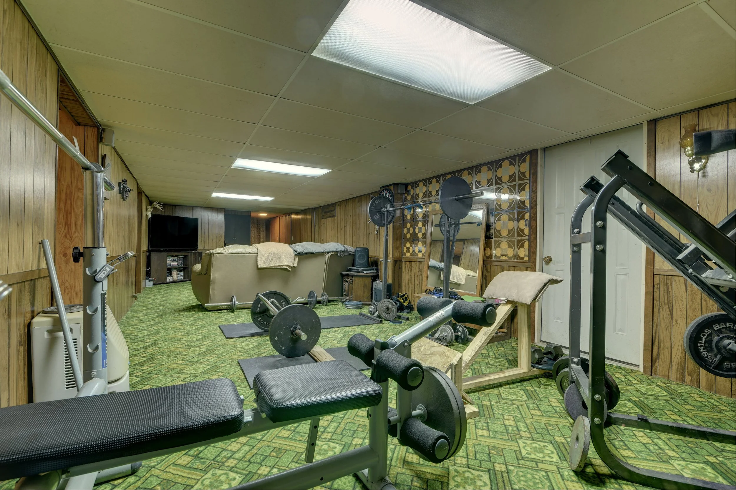 A basement gym with weightlifting equipment, a bench press, dumbbells, and a treadmill. In the background, there is a couch and a television.