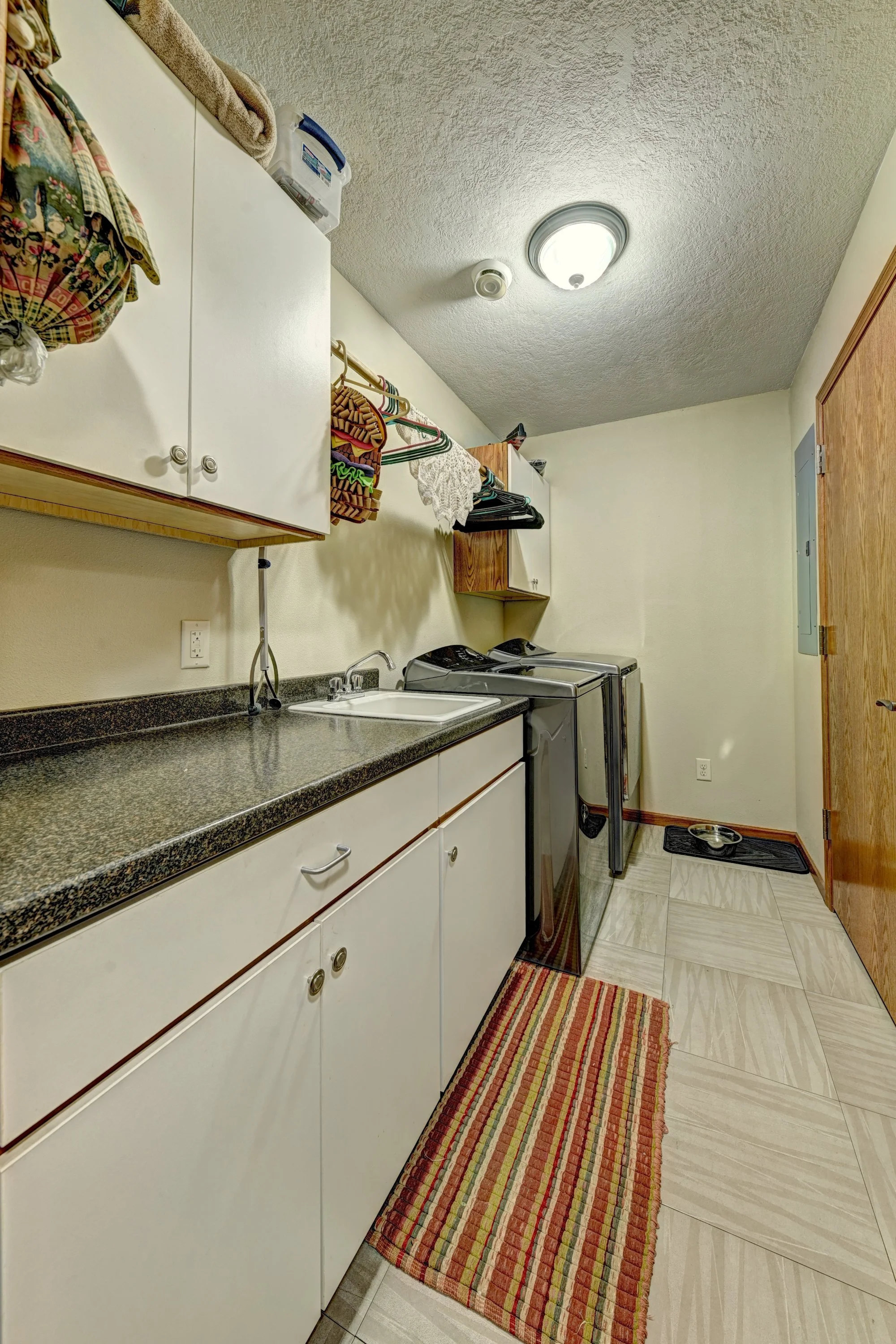 A laundry room with white cabinets, a countertop with a sink, a washing machine, and a red, yellow, and green striped rug on a tiled floor.