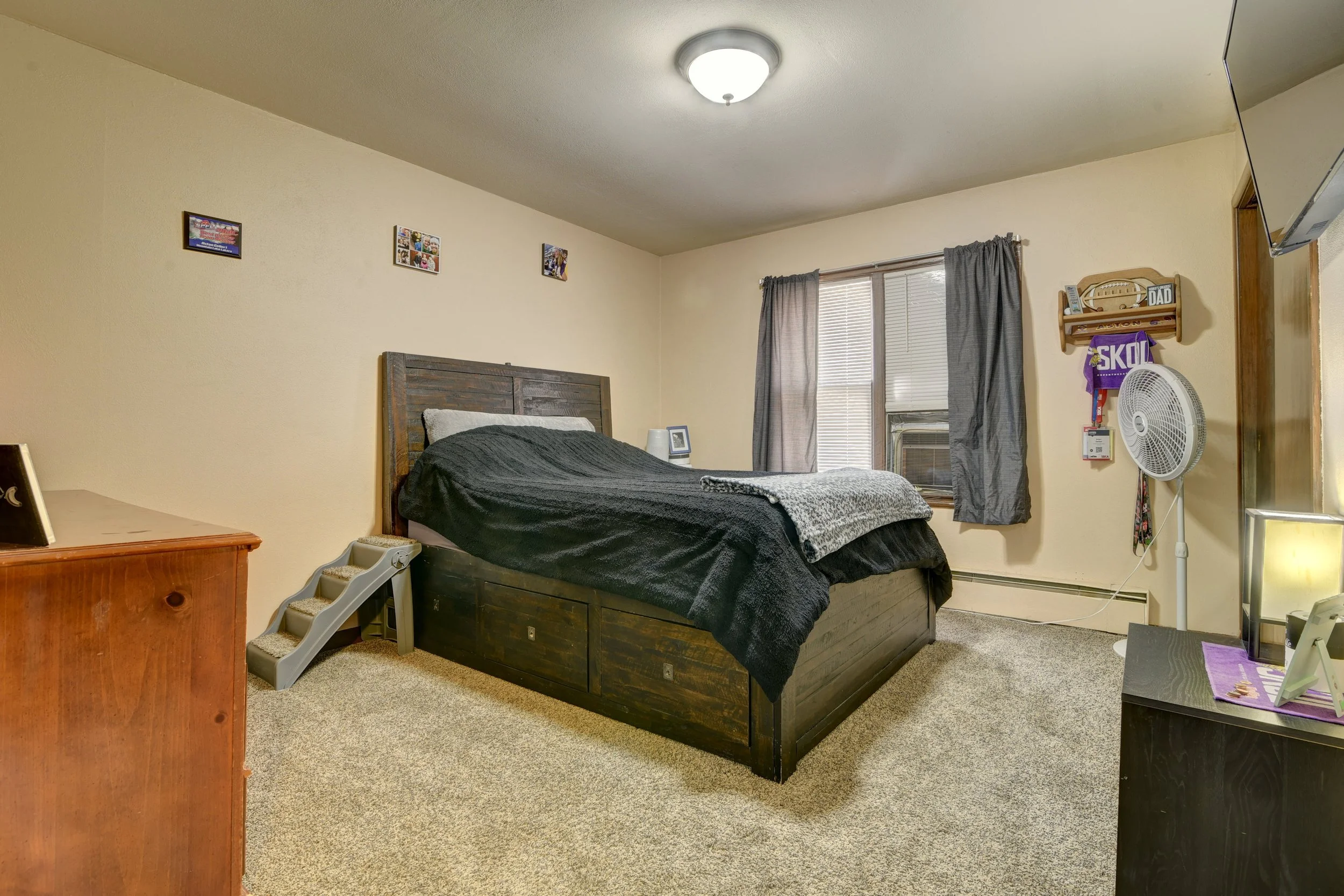 Bedroom with a bed, a window with blinds and curtains, a wooden dresser, a small step stool, a wall-mounted shelf, a standing fan, a TV, and framed photographs.